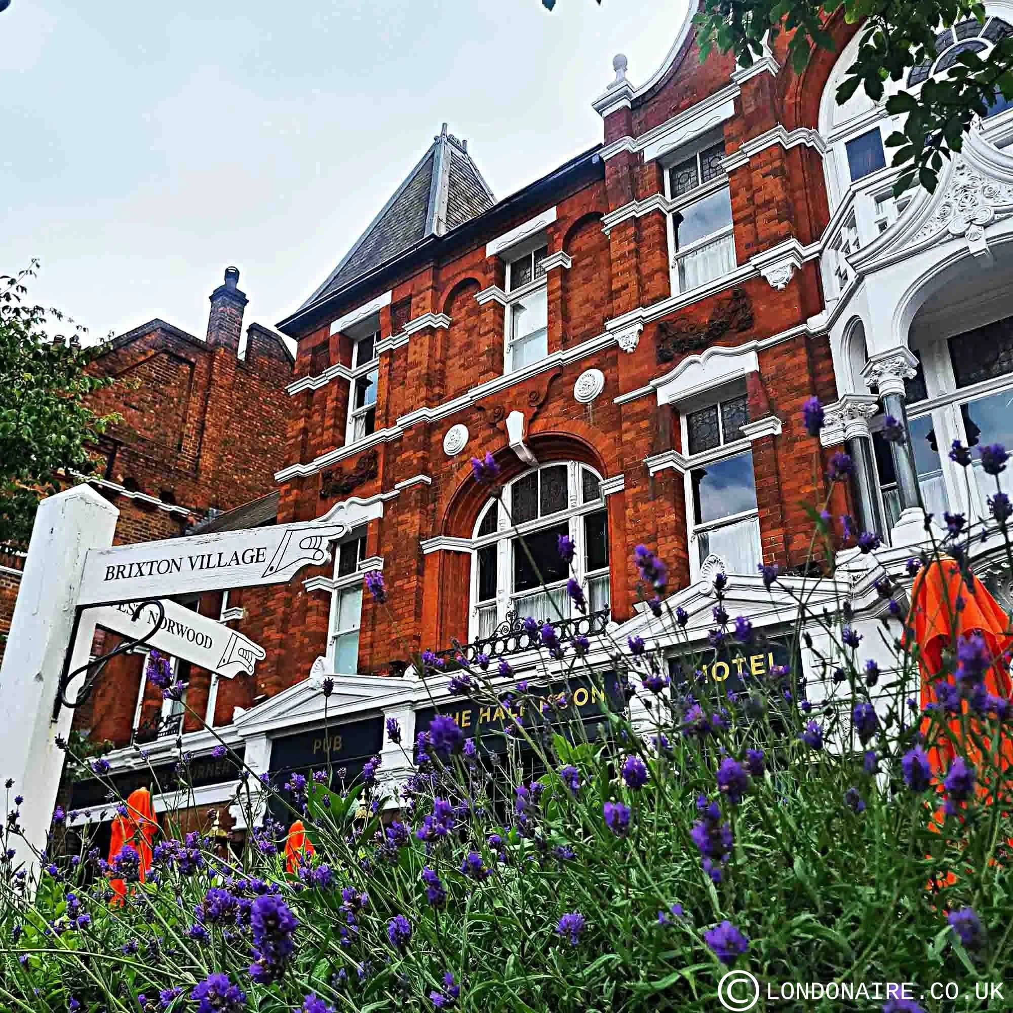 The image shows a historic red-brick building with white decorative trim, surrounded by purple flowers and green foliage, with a cloudy sky above. There is a sign indicating the location as Brixton Village in London.
