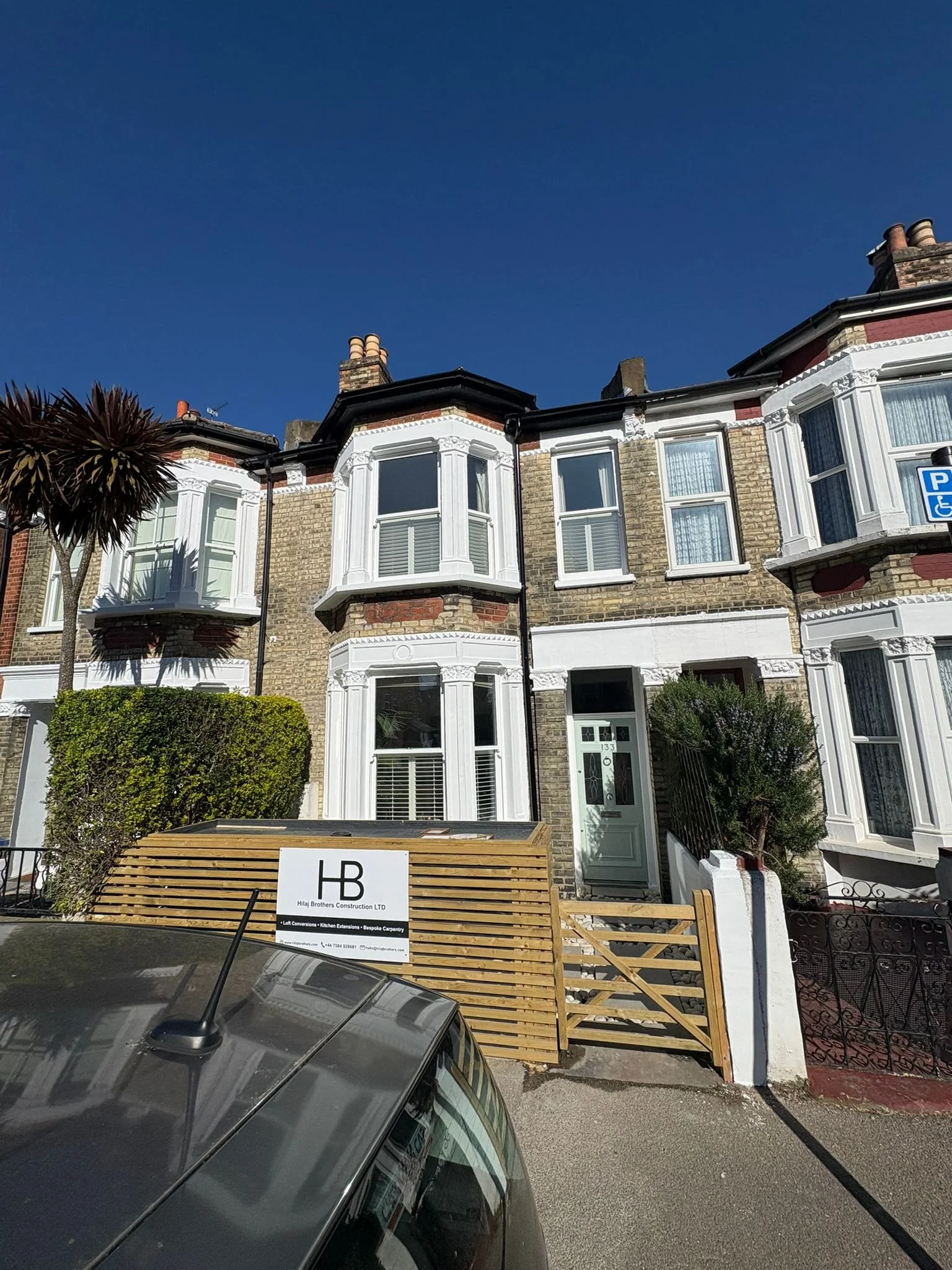 Front view of a row of Victorian-style terraced houses with bay windows and brick facades, a parking sign indicating accessible parking, a tree and shrubbery in the front garden, and a construction sign in the foreground.