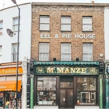 A brick building with three floors, with a sign that reads 'EEL & PIE HOUSE' on the upper level, and a storefront called 'M. MANZE' on the ground level, with a green awning and large windows.