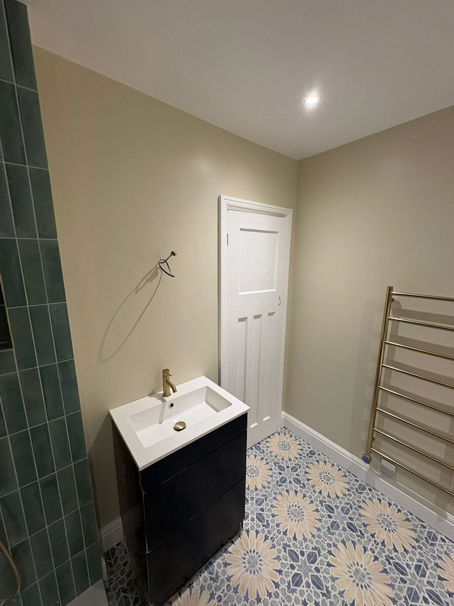 A small bathroom with beige walls, a white door, a black vanity with a white sink, a gold faucet, a patterned tile floor, and a towel rack on the wall.