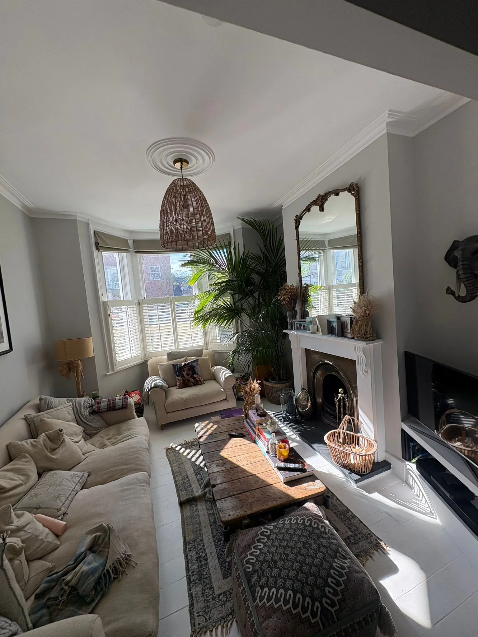 Living room with beige sofas, a wooden coffee table, a fireplace, plants, framed pictures, and a TV. Sunlight streams through window blinds.
