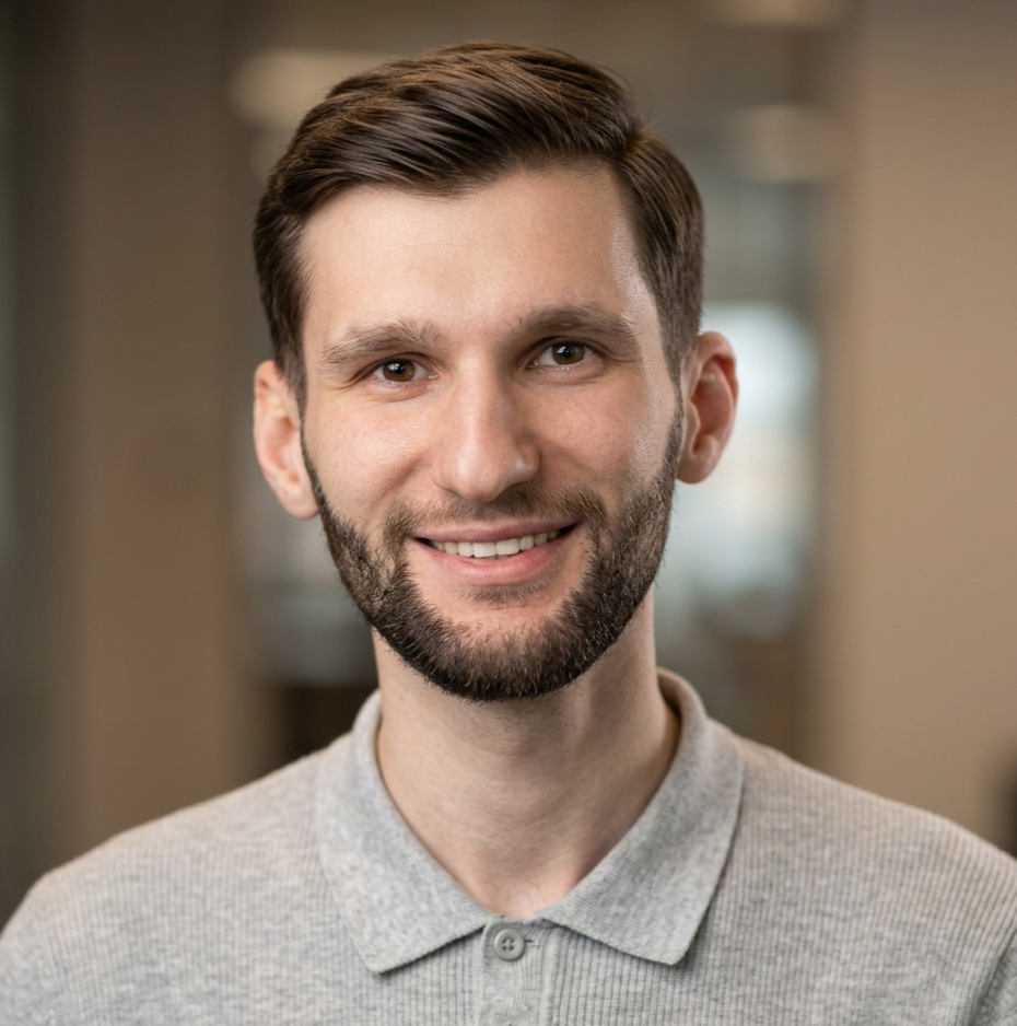 Portrait of a young man with short brown hair and a beard, smiling, wearing a light gray collared shirt, indoors with blurred background.