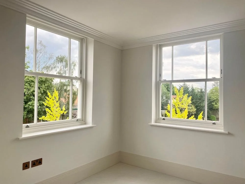 Empty white room with two large windows showing trees outside and a partly cloudy sky.