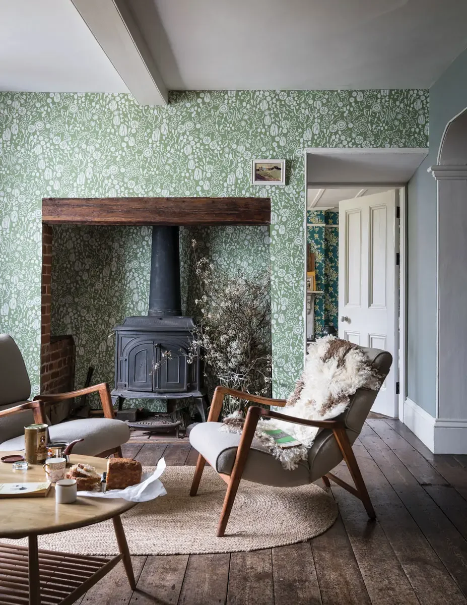 A cozy living room with vintage furniture, a wood-burning stove, and green floral wallpaper. There are two mid-century modern chairs, a round wooden coffee table with snacks and cups, a round beige rug, and a white door leading to another room. Natural light brightens the space.