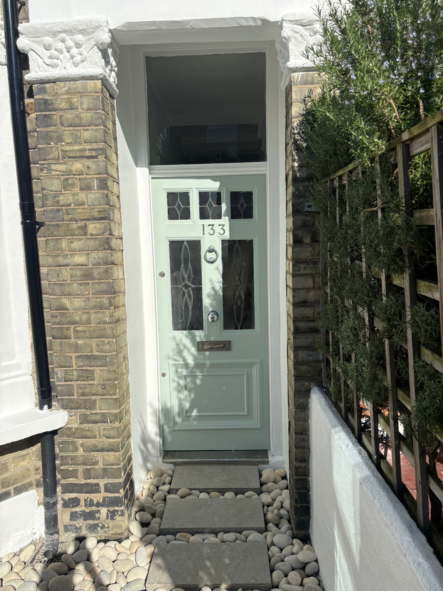 Front porch of a house with a pale green door featuring decorative glass panels, house number 133, and a white frame, surrounded by brick columns, greenery on the right, a pathway of rectangular stepping stones, and white rocks on either side.