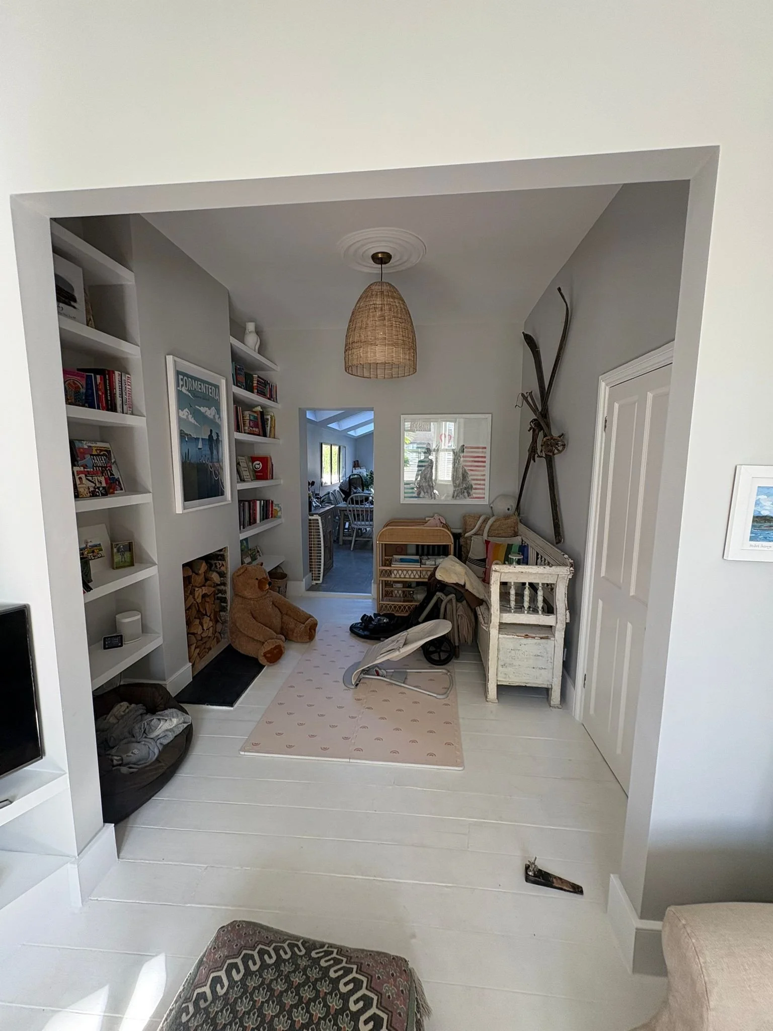 Living room interior with white walls, a ceiling light with a woven lampshade, a built-in bookshelf filled with books and decorative items, a window with two mannequin busts, a white distressed storage chest, a toy stroller, a teddy bear, and scatter