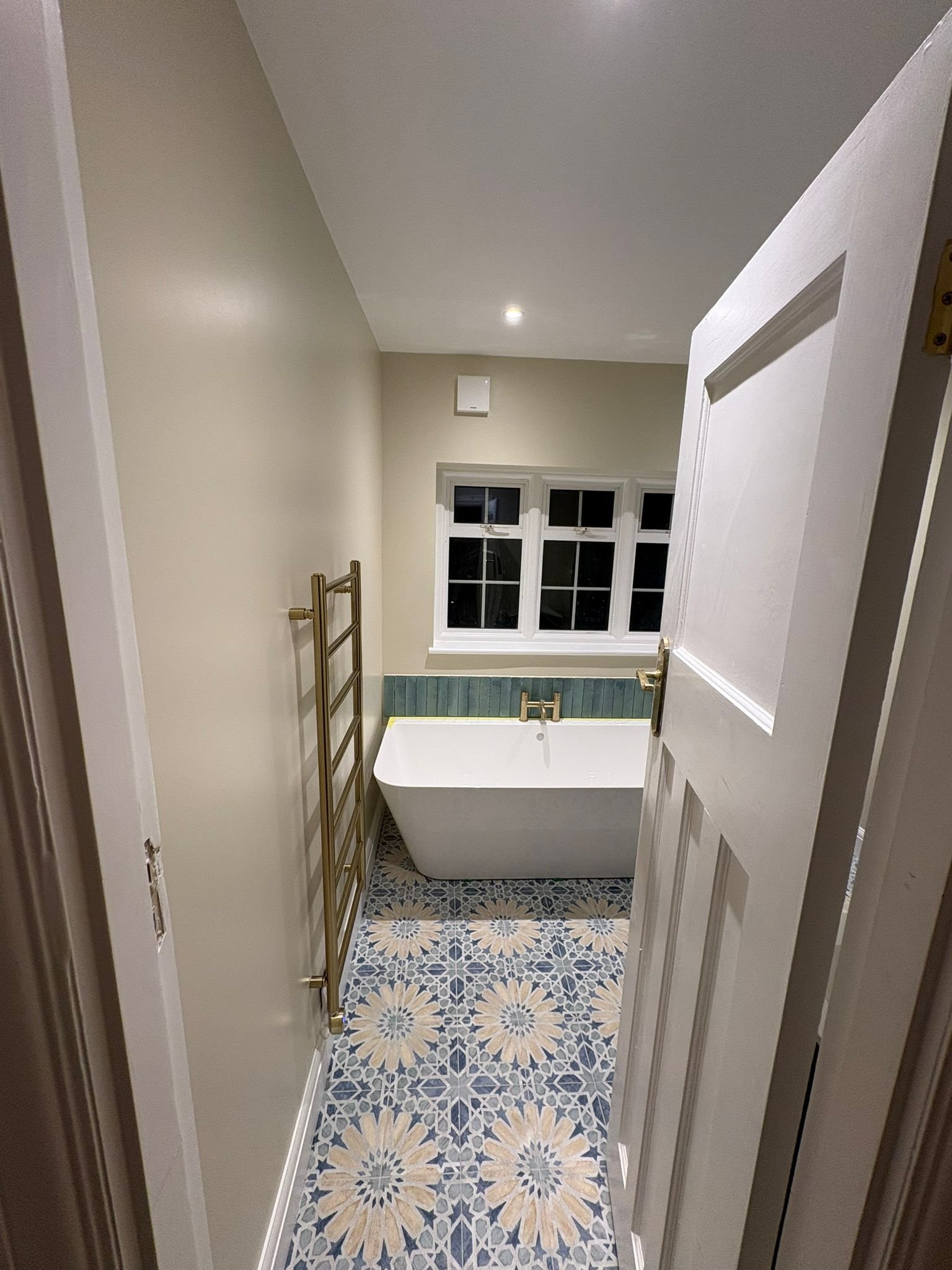 View of a small bathroom with patterned tile floor, a white bathtub, a window with four panes, a gold towel rail, and neutral-colored walls.