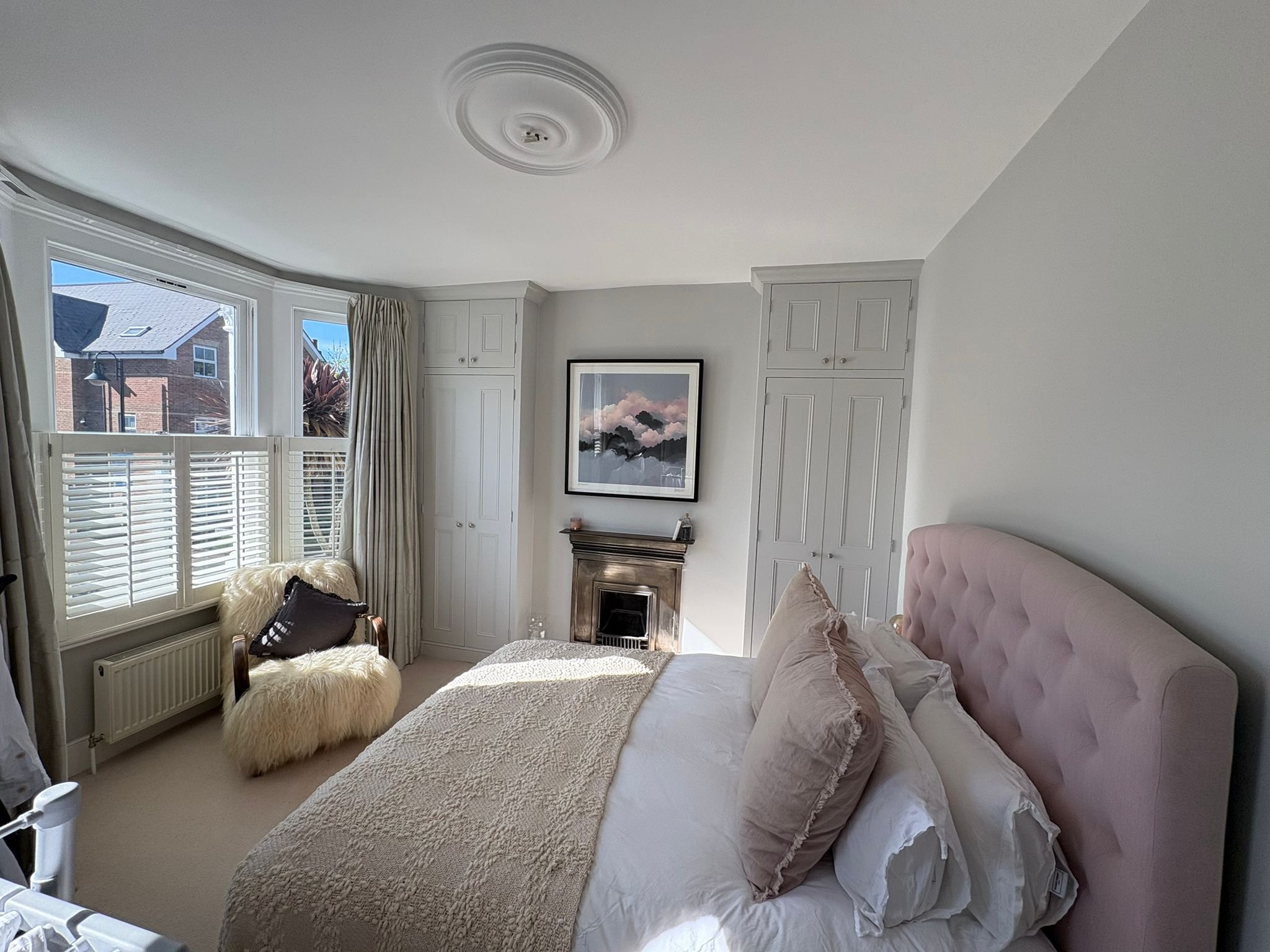 A cozy bedroom with a large bed with a pink tufted headboard, beige pillows, and a textured beige throw blanket. There is a fluffy beige ottoman at the foot of the bed. The room features white walls, built-in cabinets, and a fireplace with a framed l
