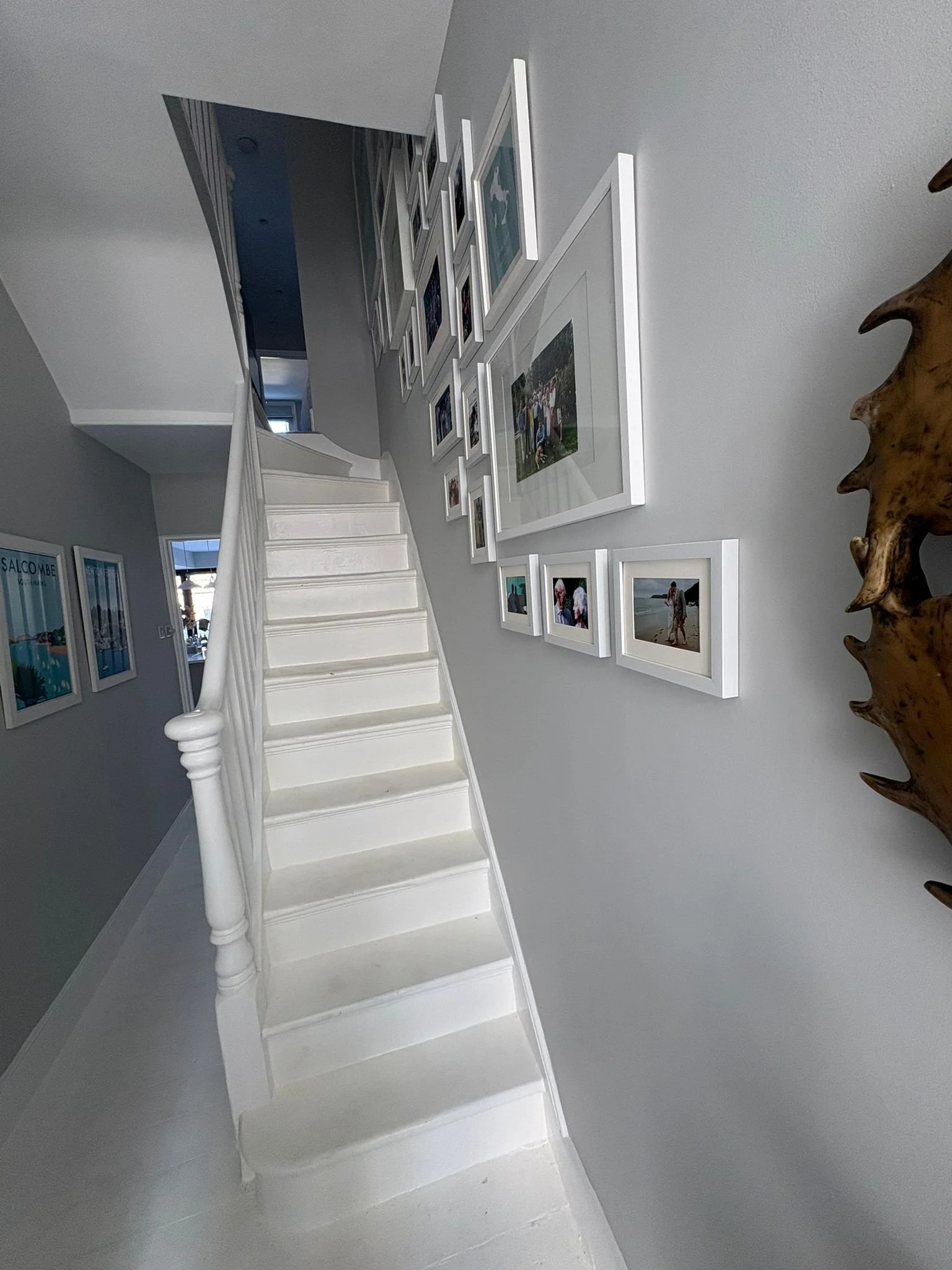 Interior view of a staircase in a home, with white stairs, a gallery wall of framed photos, and a wooden decorative piece on the right.
