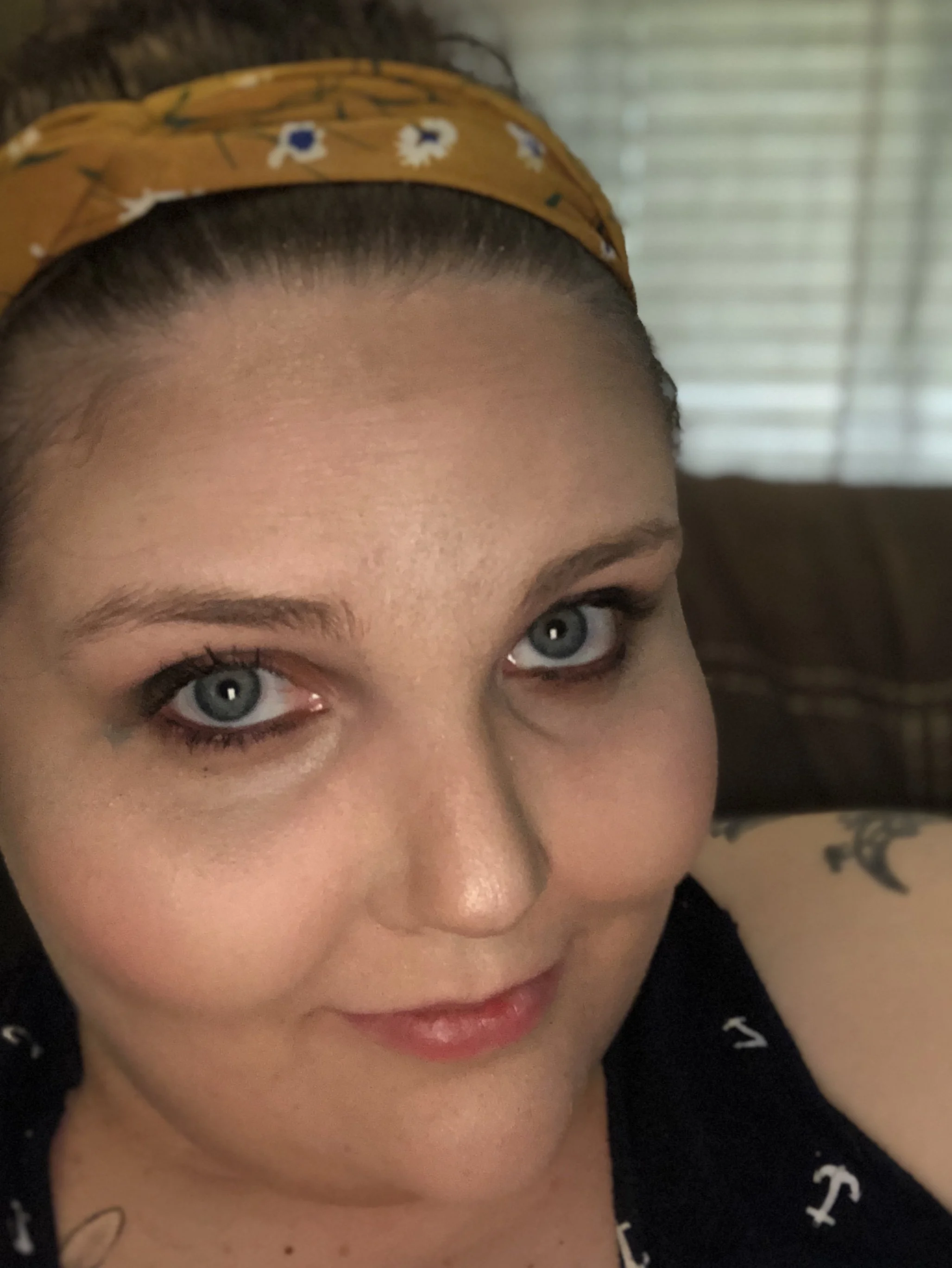 Close-up of a woman with blue eyes, wearing a yellow headband with a floral pattern, black top with white nautical symbols, and a small tattoo on her shoulder, sitting indoors with a window and blinds in the background.