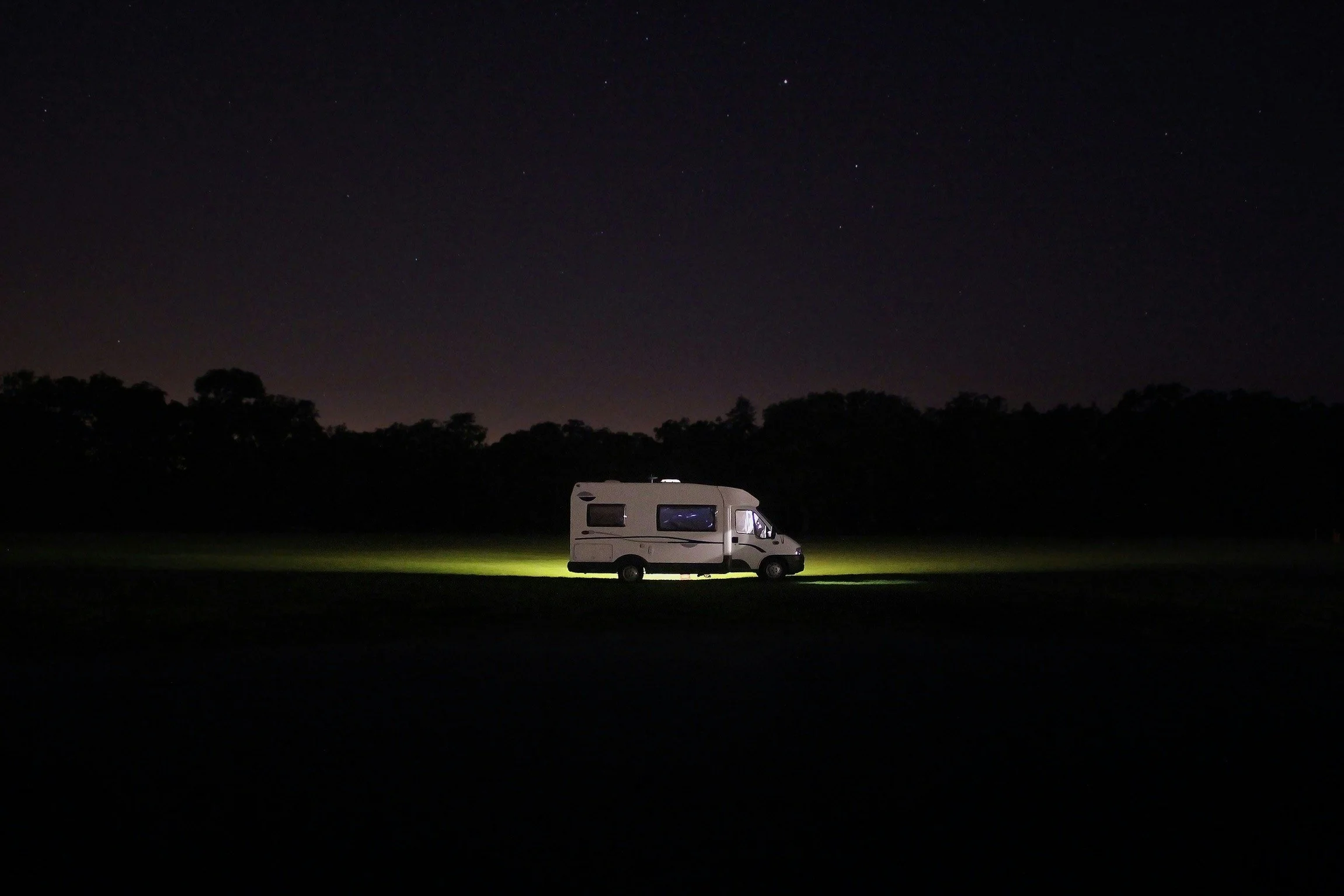 A white camper van parked on a grassy field at night under a starry sky, with a line of trees in the background.