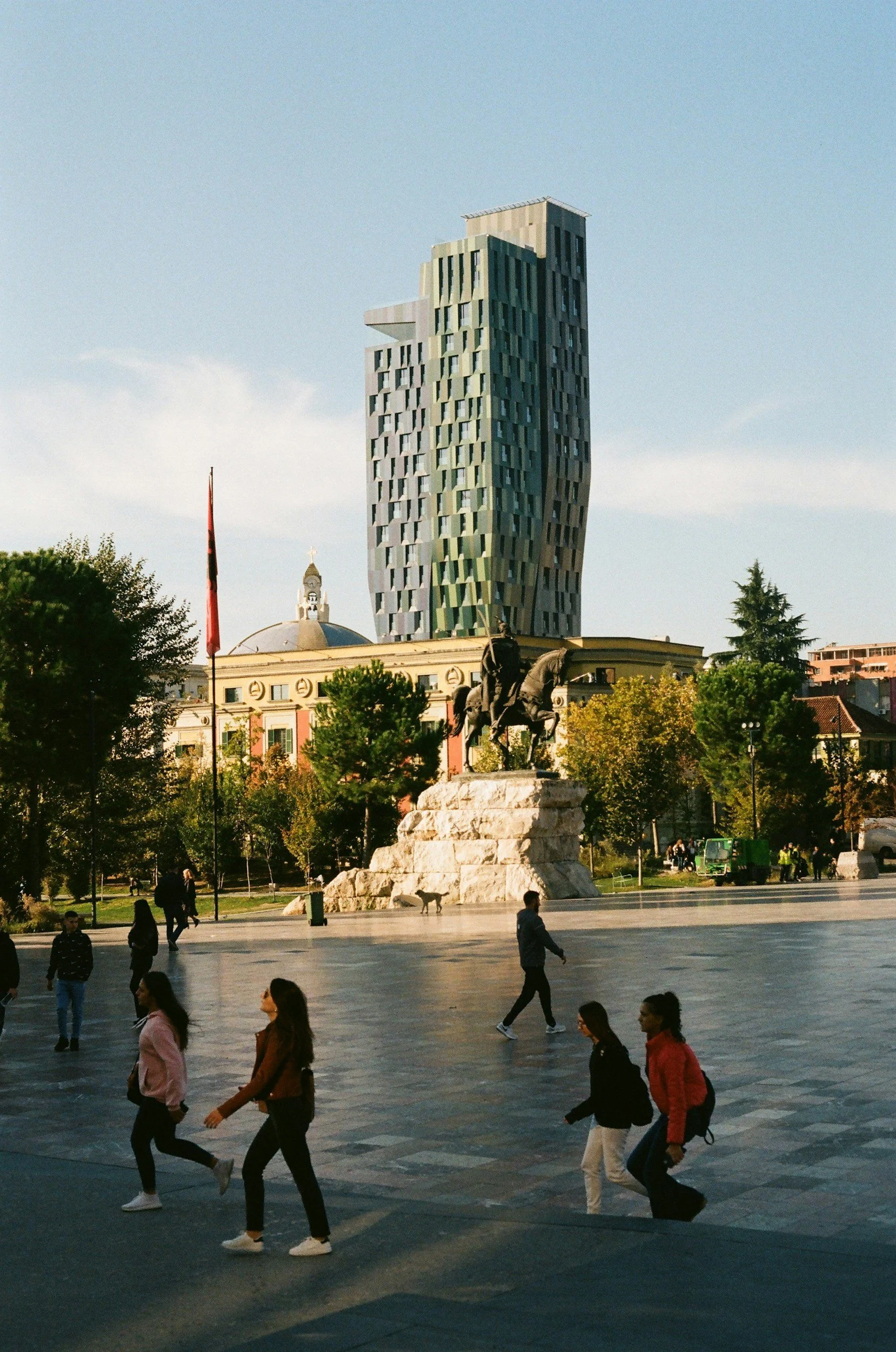 People walking in a city square with a historic statue in the foreground and a modern twisting skyscraper in the background.