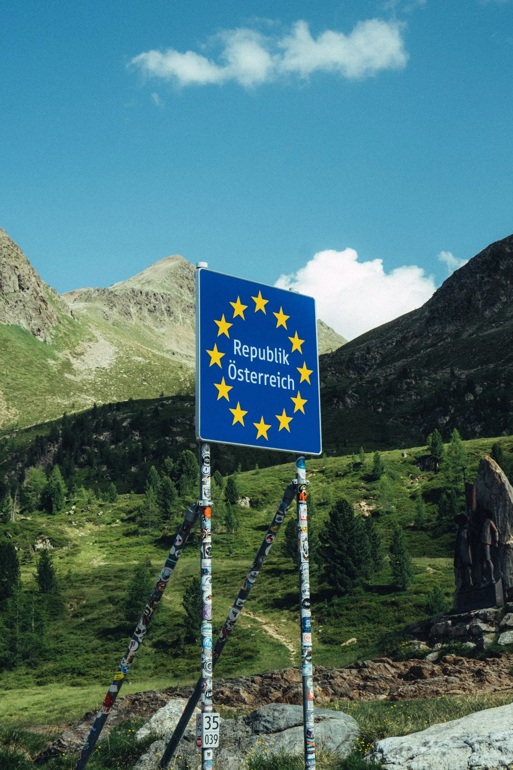 A blue sign with yellow stars and the words 'Republik Österreich' stands amidst a green mountainous landscape under a partly cloudy sky.