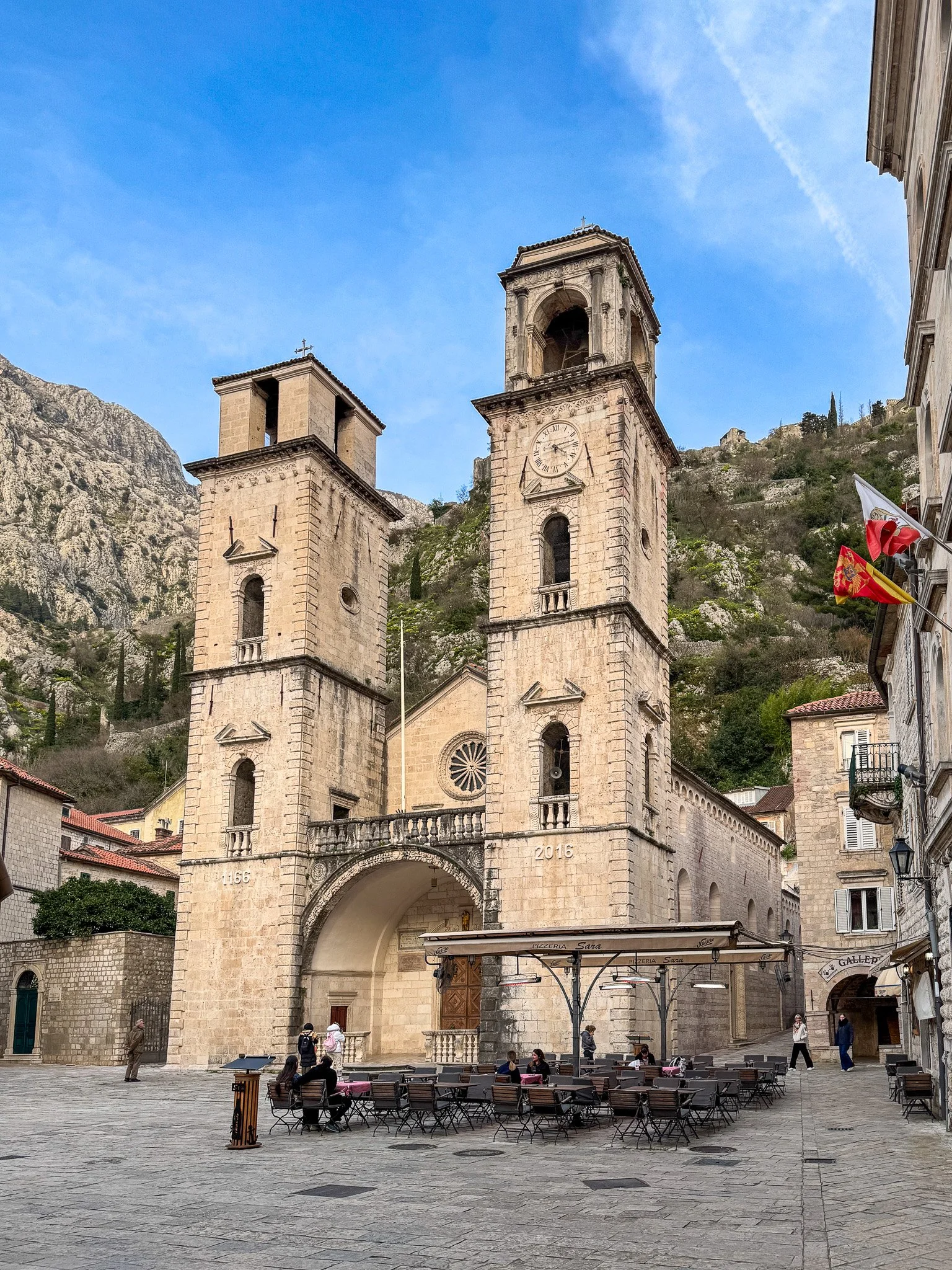 The west face of the Cathedral of Saint Tryphon in Kotor, Montenegro, on either side, situated in a town square with outdoor seating and people walking or sitting. Flags are visible on the right side.