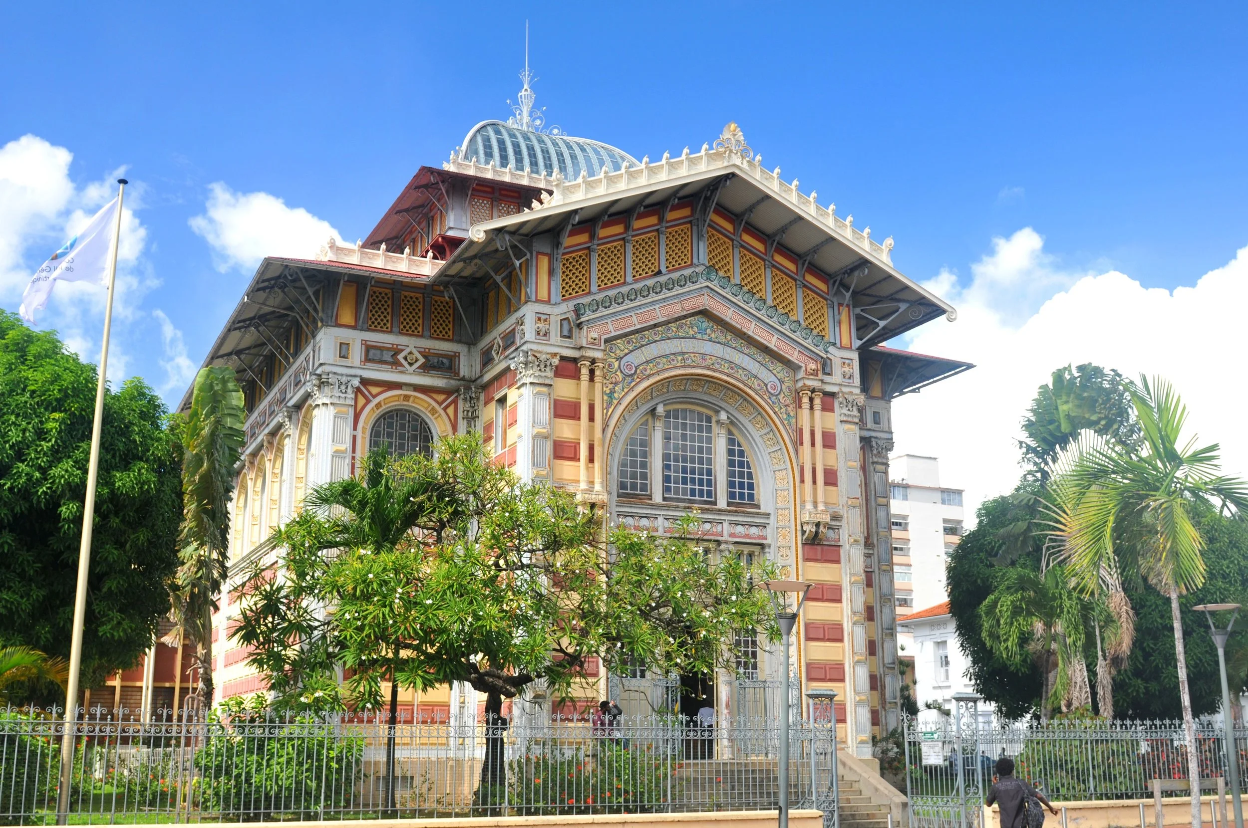 A historic ornate building with intricate architectural details, colorful facade, large arched windows, and decorative elements, surrounded by lush green trees and a fence, under a blue sky with clouds.
