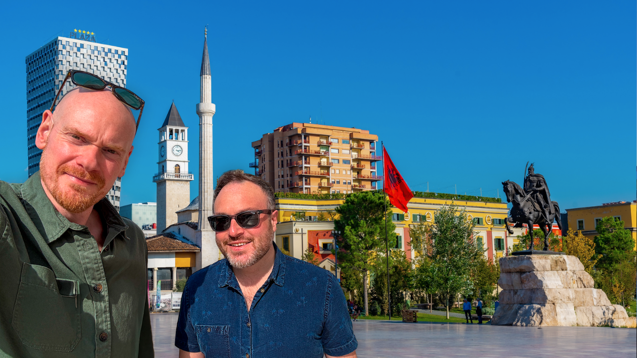 Two men taking a selfie in Skanderbeg Square, Tirana, Albania, with a statue of a mounted warrior, a red flag, a clock tower, and modern buildings in the background.