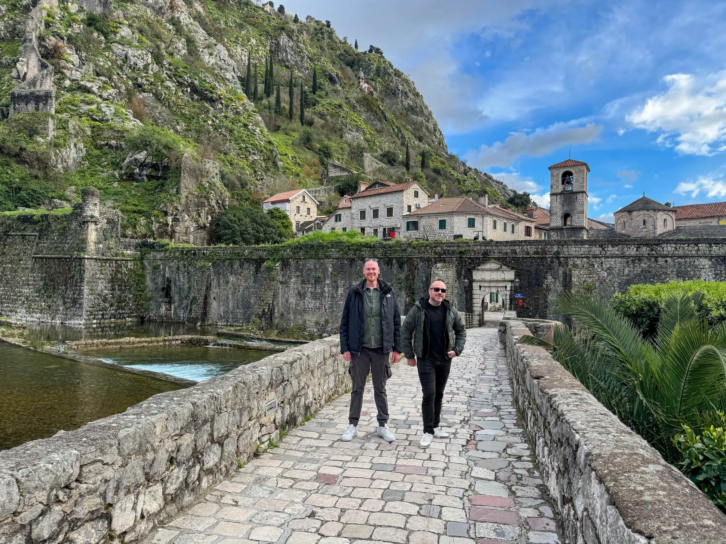 Three men walking on a cobblestone path alongside a river in Kotor, Montenego with stone buildings and a church with a bell tower in a scenic mountainous area under a partly cloudy sky.