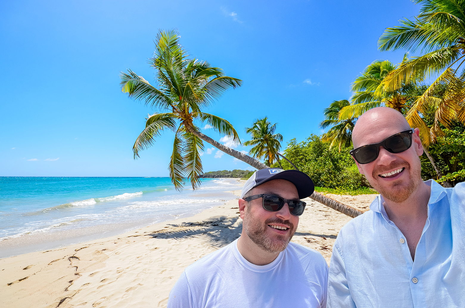 Two men smiling on a sunny beach with palm trees, ocean waves, and clear blue sky in the background.