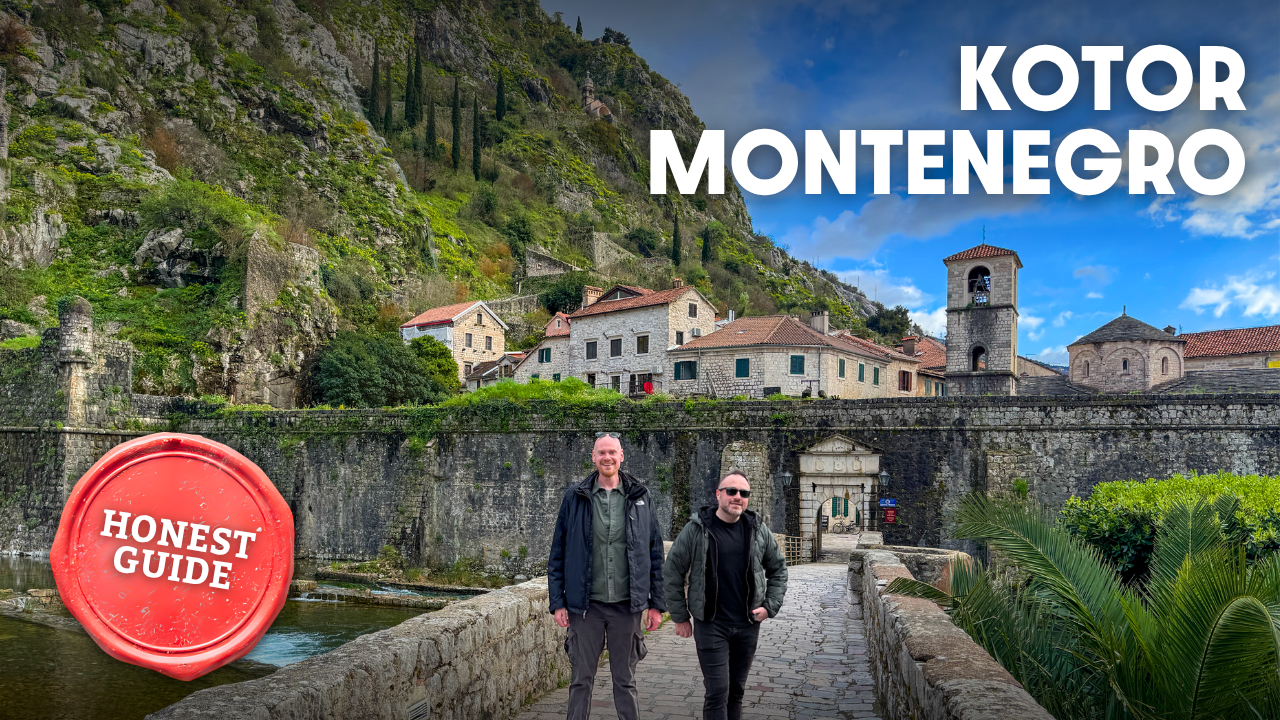 Two men walking on a stone bridge with historic buildings and a hillside in Kotor, Montenegro, under a partly cloudy sky.