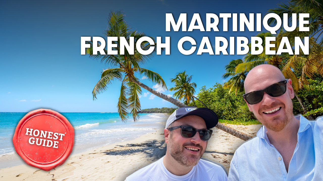 A sunny Caribbean beach scene with two men smiling, palm trees, and clear blue water, featuring text 'Martinique French Caribbean' and a red stamp-like icon reading 'Honest Guide'.