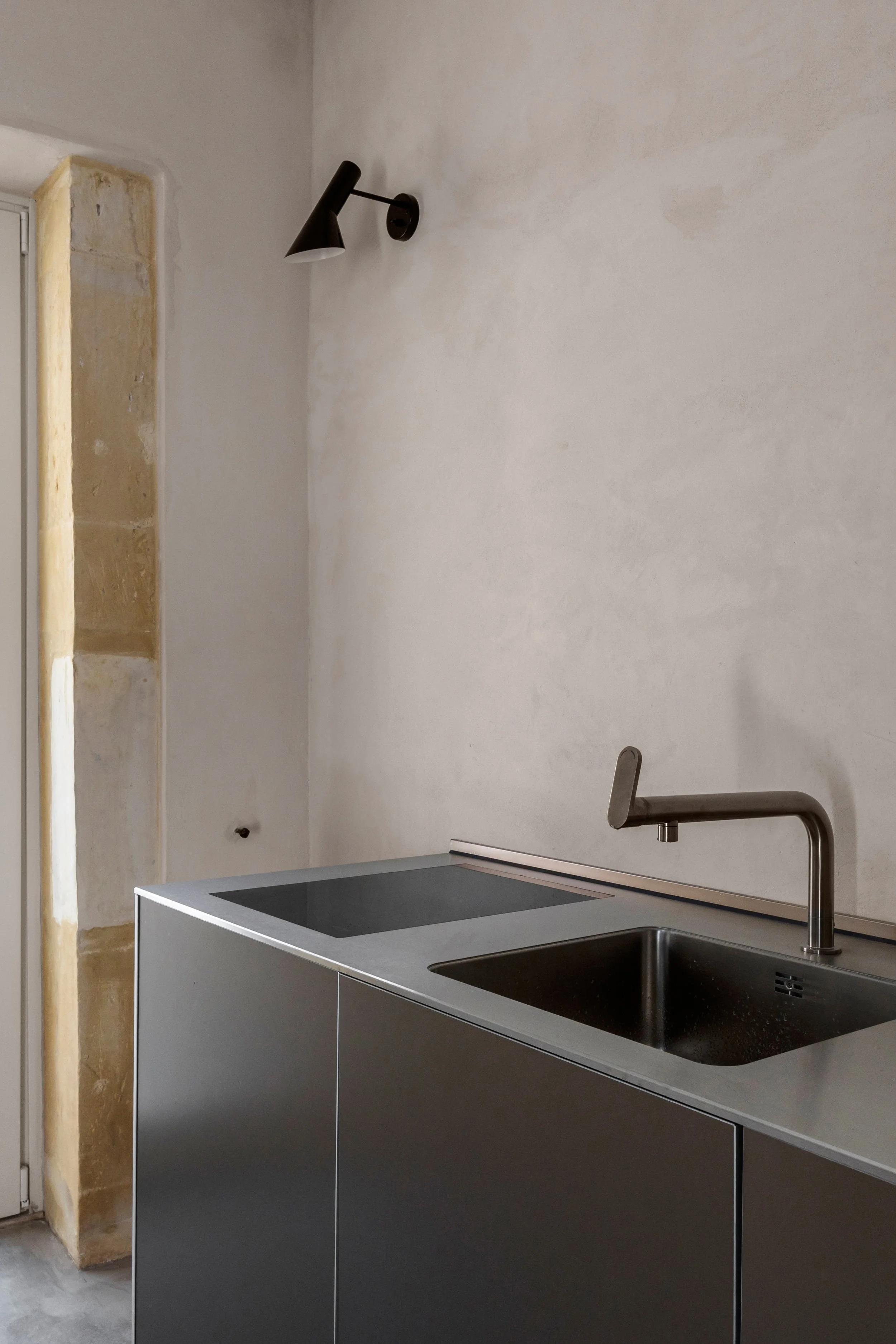 Modern kitchen with a stainless steel sink and faucet, black stovetop, and a wall-mounted black lamp.