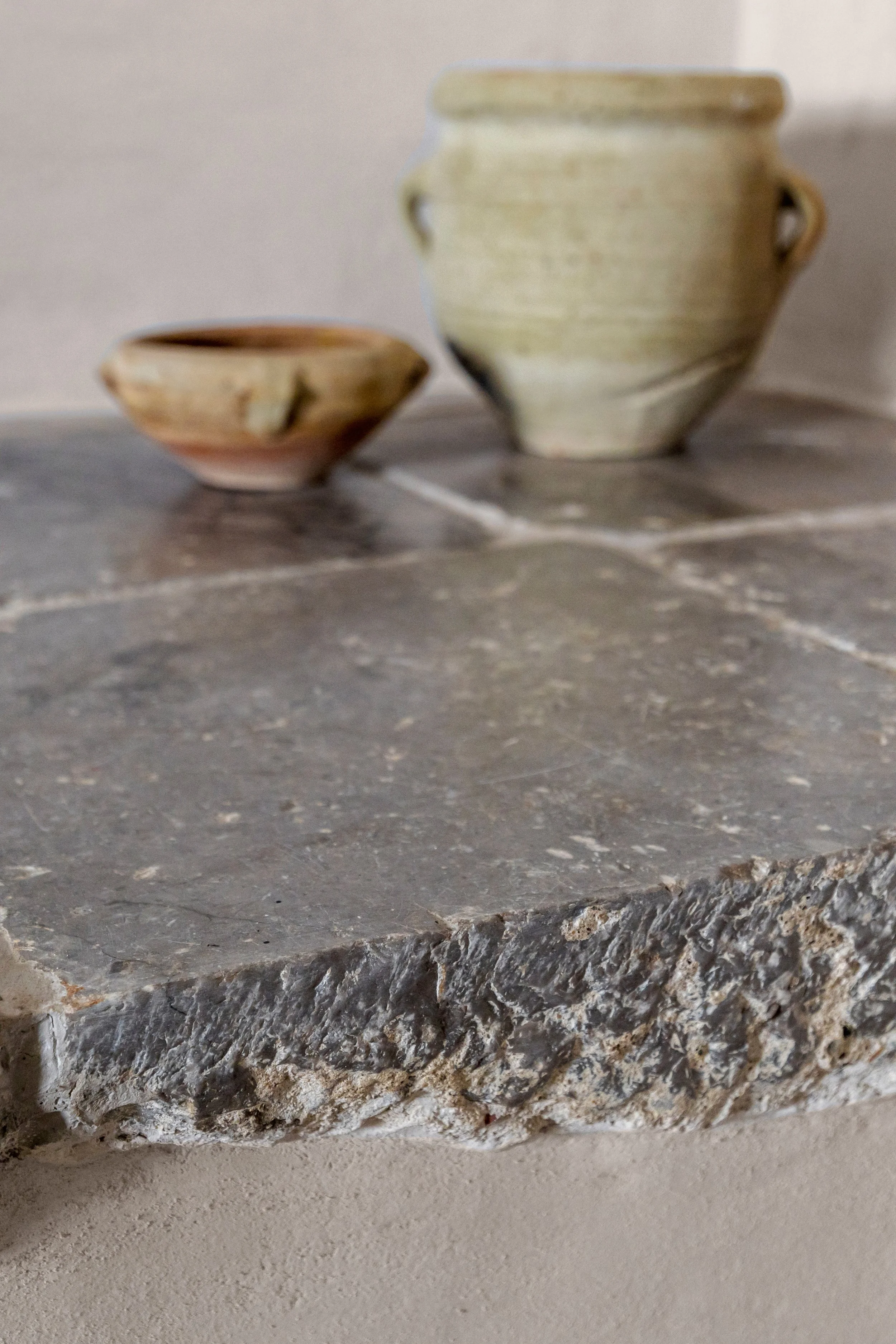 Close-up of a rough, textured stone countertop with two pottery vessels in the background, one small and one large.