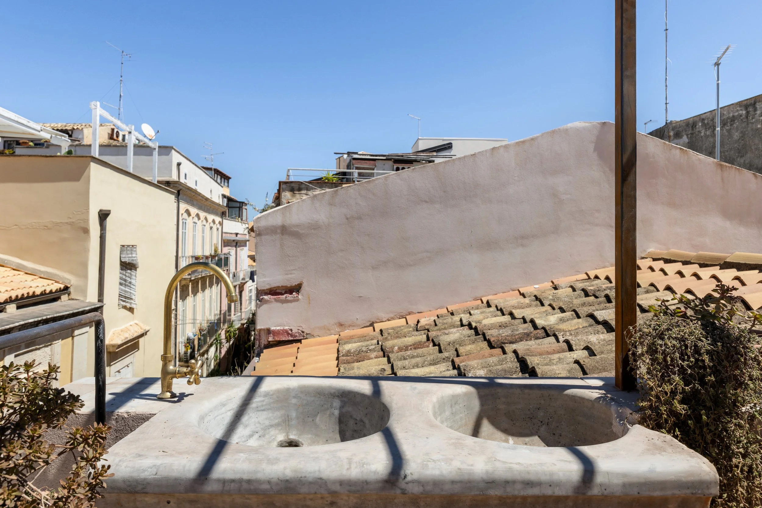 An outdoor double sink with a brass faucet on a concrete countertop, overlooking a cityscape with various buildings, rooftops, antennas, and plants, under a clear blue sky.