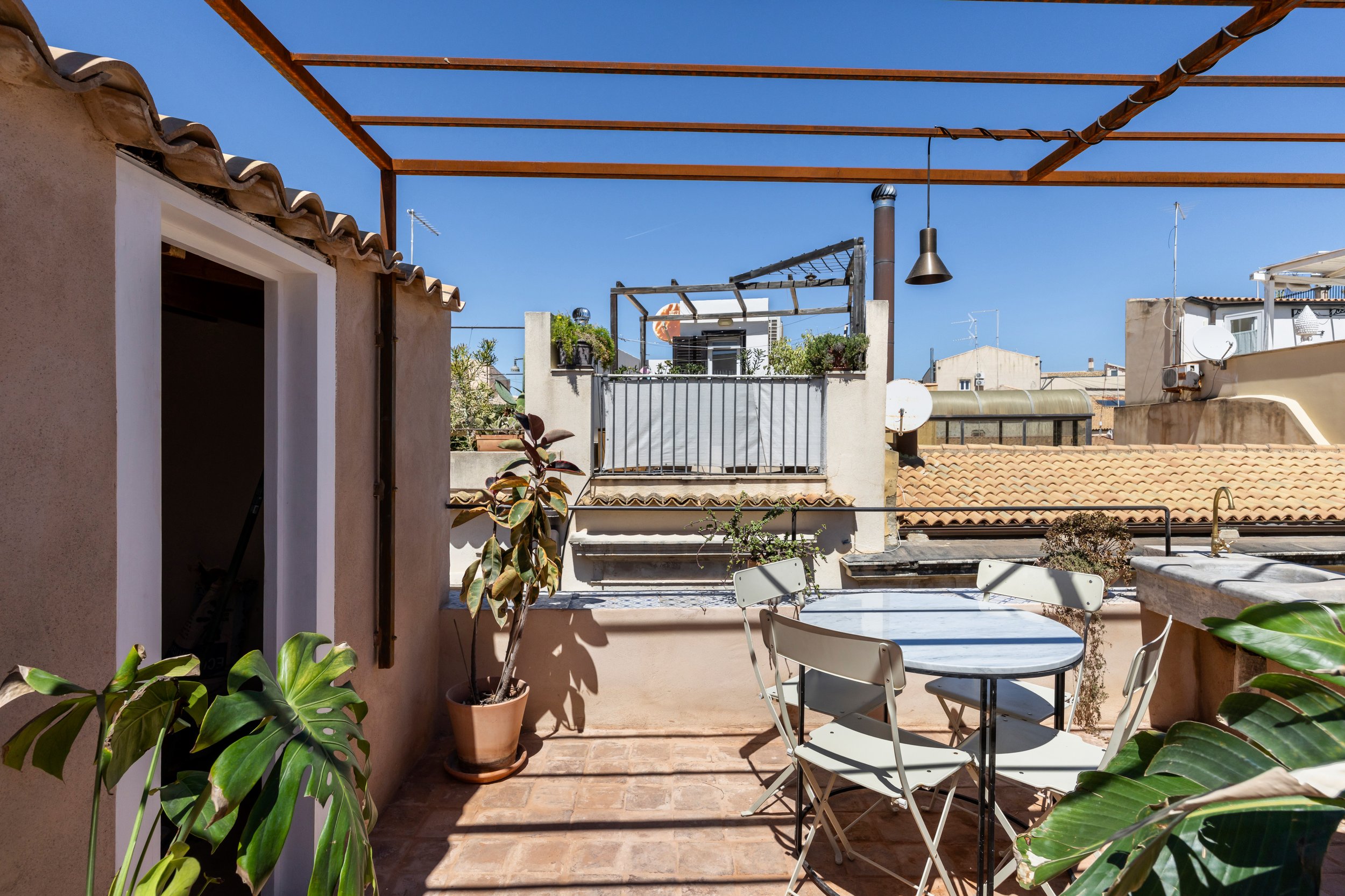 Rooftop patio with a round table and four chairs, potted plants, and a view of neighboring buildings with tiled roofs under a clear blue sky.