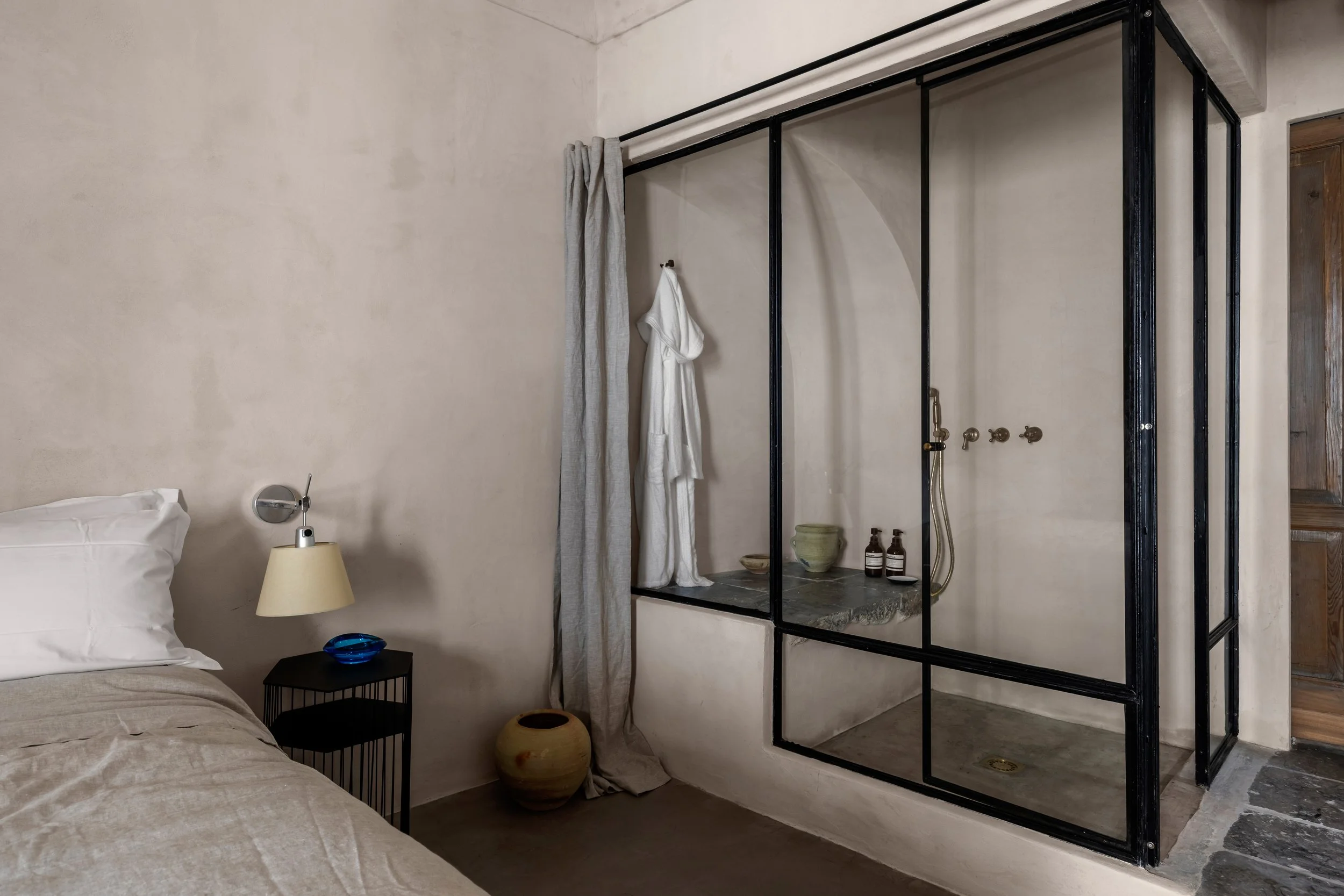Bedroom with beige walls, a bed with white pillows, a small black nightstand with a beige lamp and blue bowl, and a glass shower enclosure with black frame, a robe hanging outside, and shampoo bottles inside.