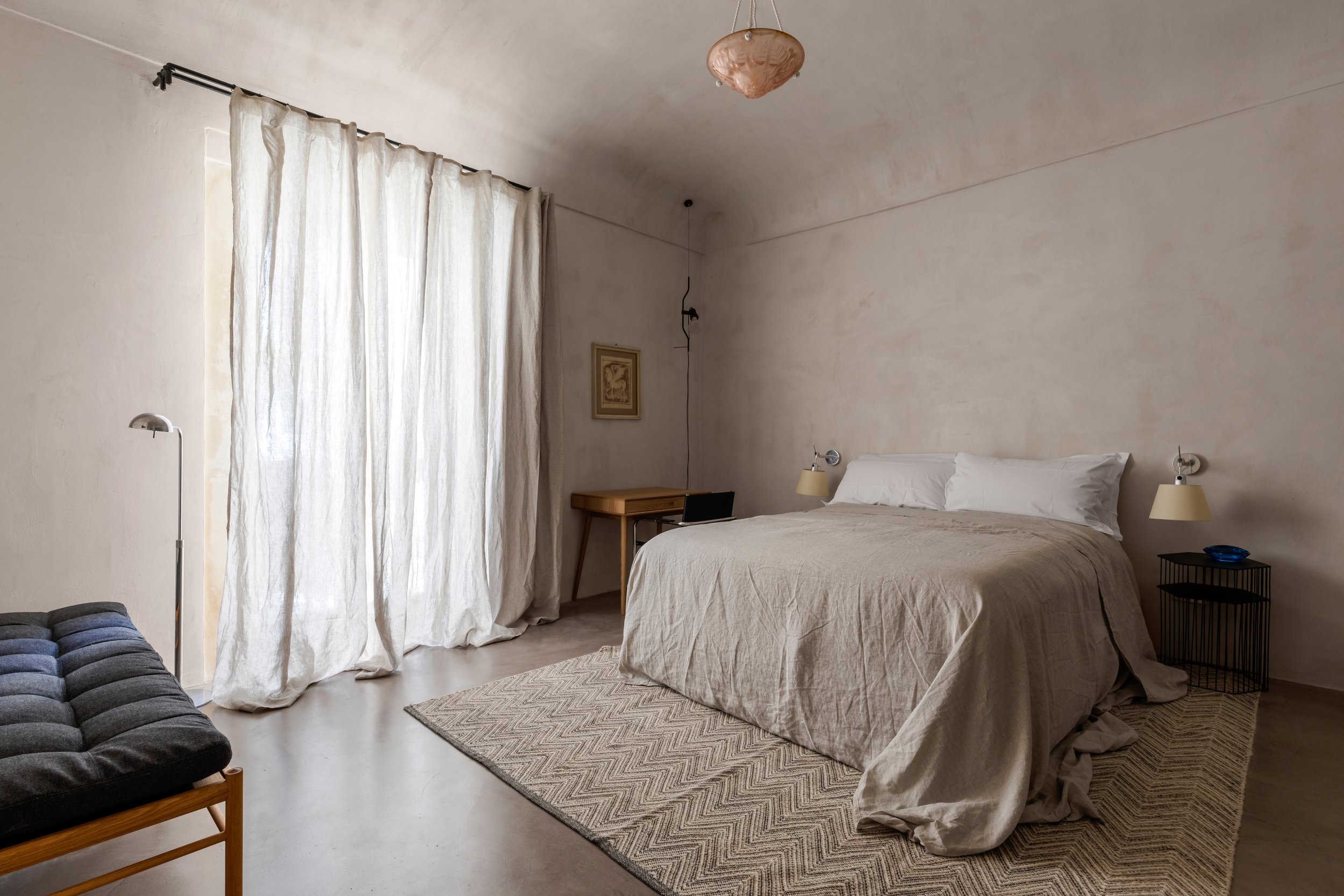 Minimalist bedroom with a beige bed, two bedside lamps, a black side table, a wooden desk, a framed picture on the wall, beige curtains, a floor lamp, and a blue bowl on the side table.