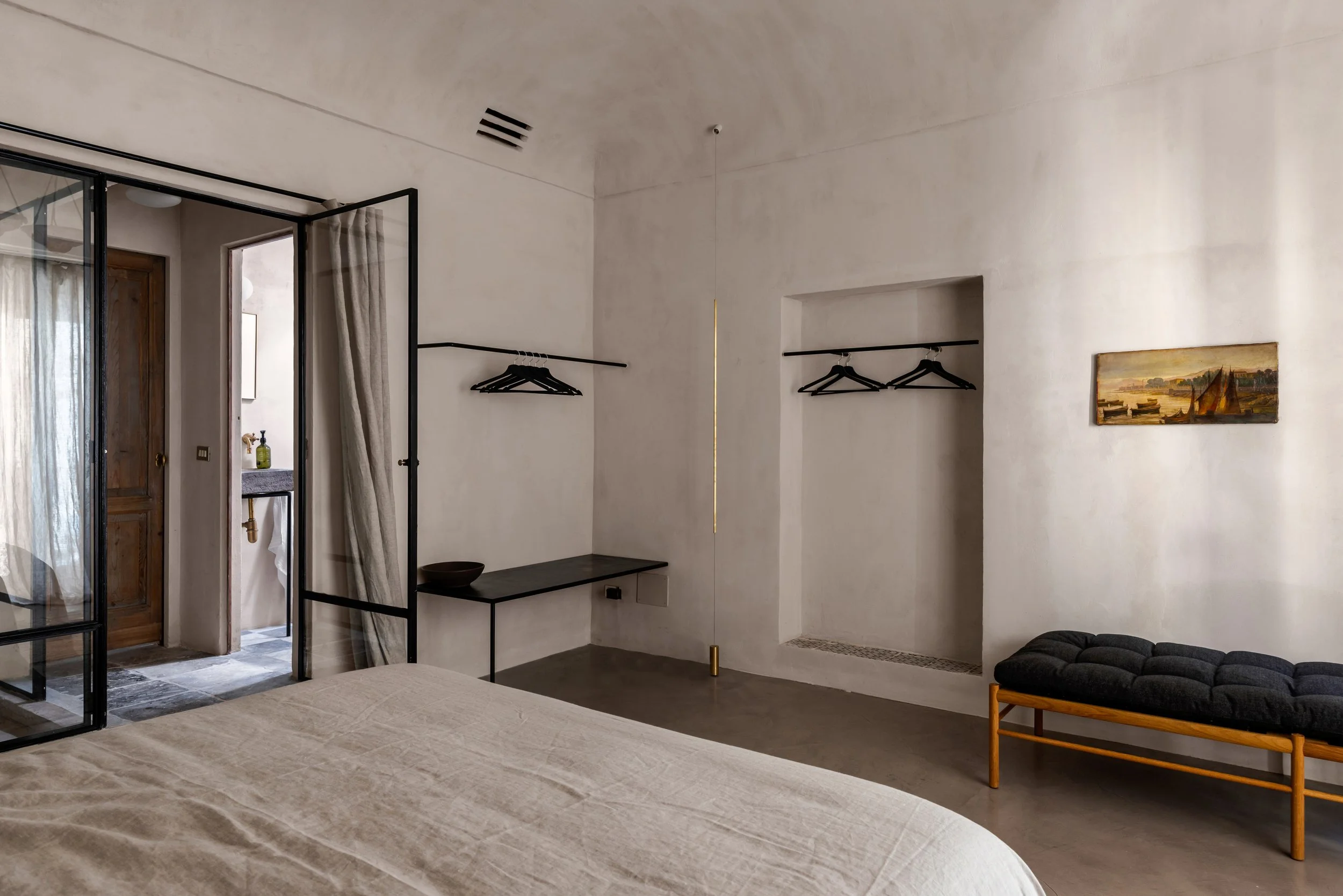 Minimalist bedroom with a bed, a small seating bench, open closet with hangers, and a partial view of an en-suite bathroom.