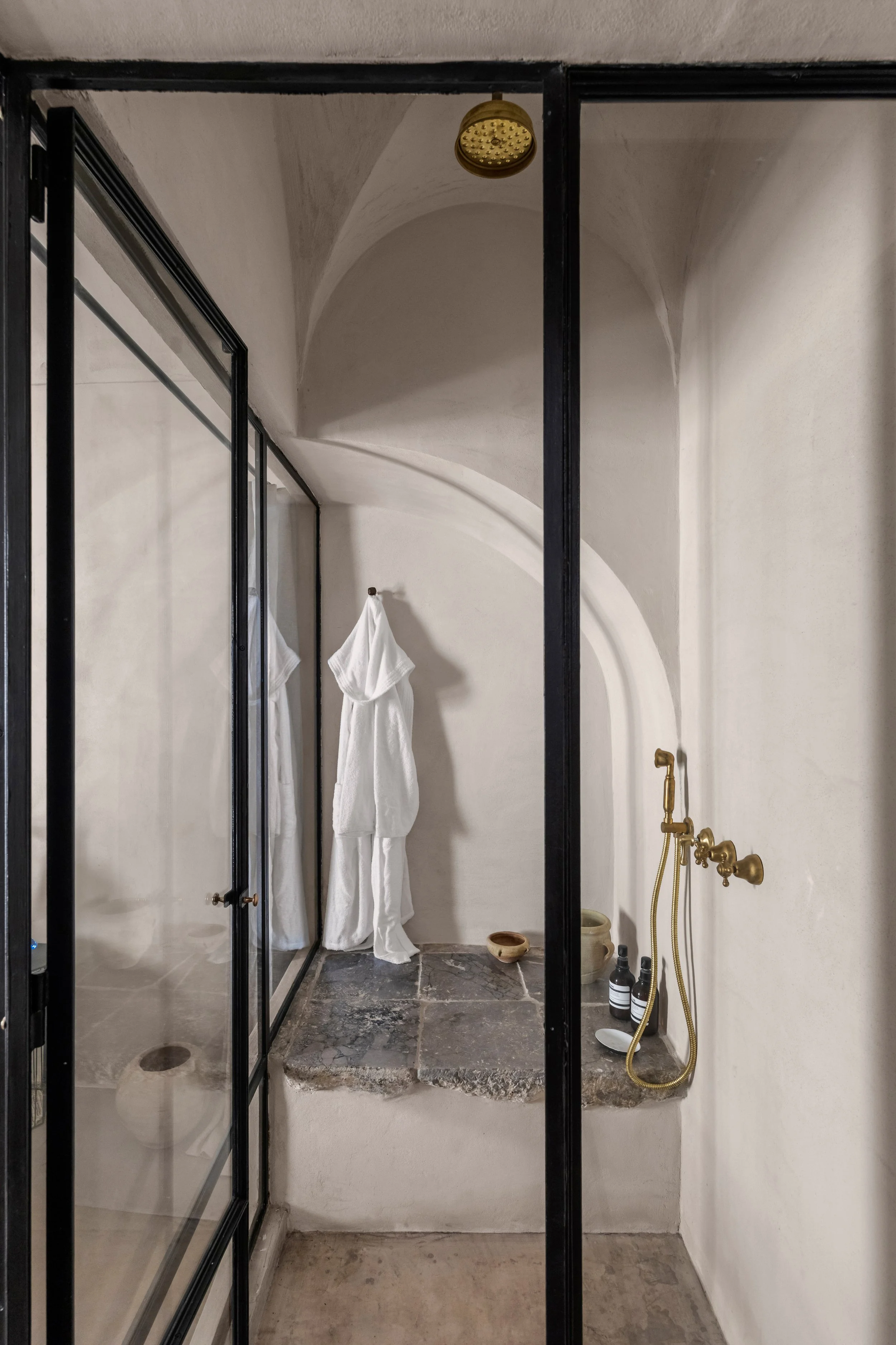 A modern shower with a glass door, a brass showerhead, and wall-mounted brass fixtures. A white bathrobe hangs on a hook on the wall. The shower area has a stone platform with toiletries, and the walls and ceiling are smooth and curved.