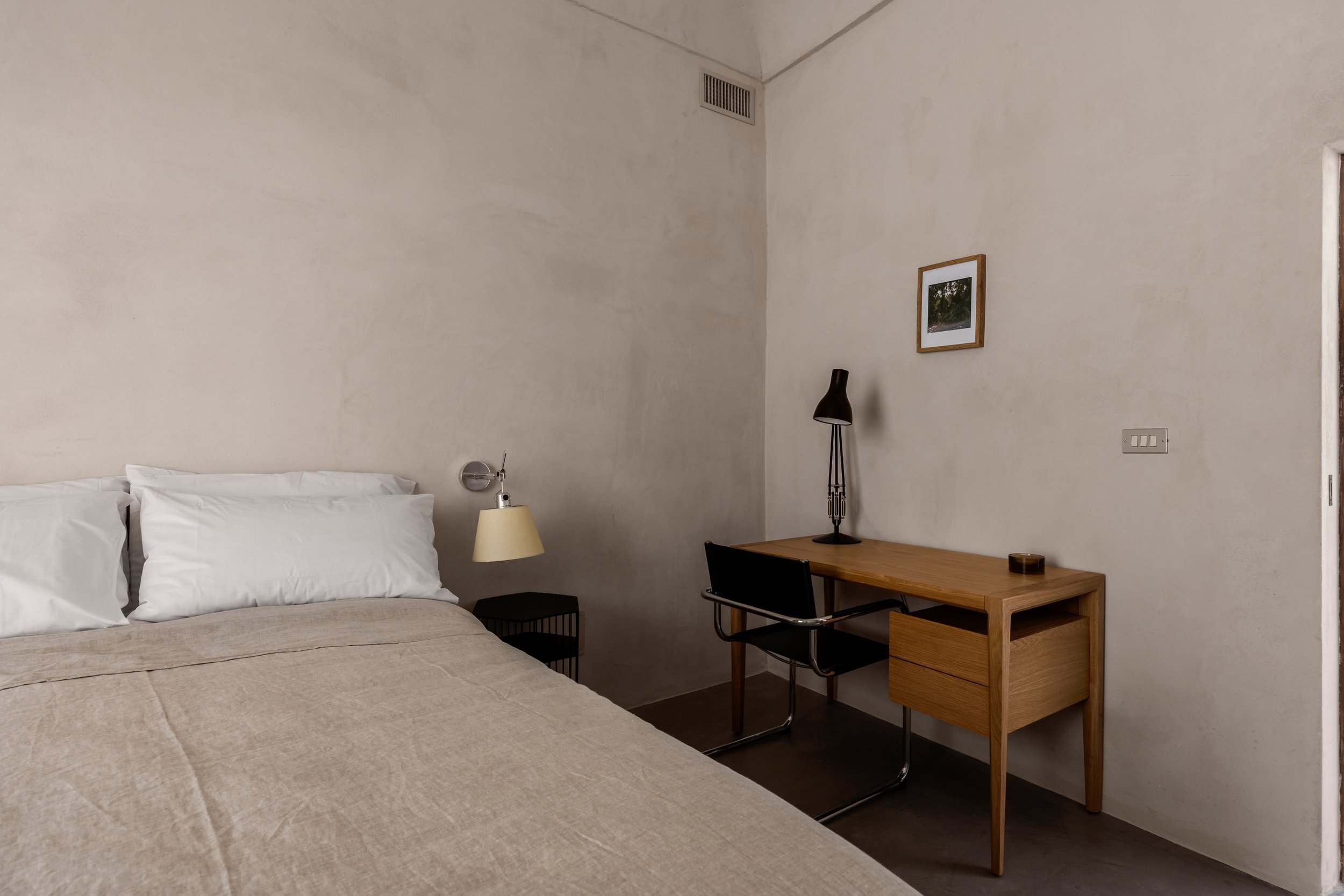Simple bedroom with a bed, bedside table with lamp, desk, chair, and wall frame.