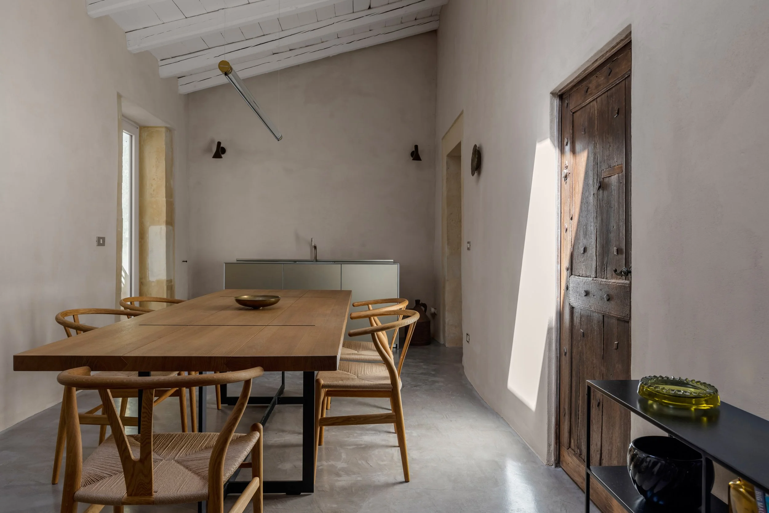 Minimalist dining room with a wooden table and six chairs, white plaster walls, a wooden door, and a black metal shelf with decorative items. Sunlight casts shadows on the wall.