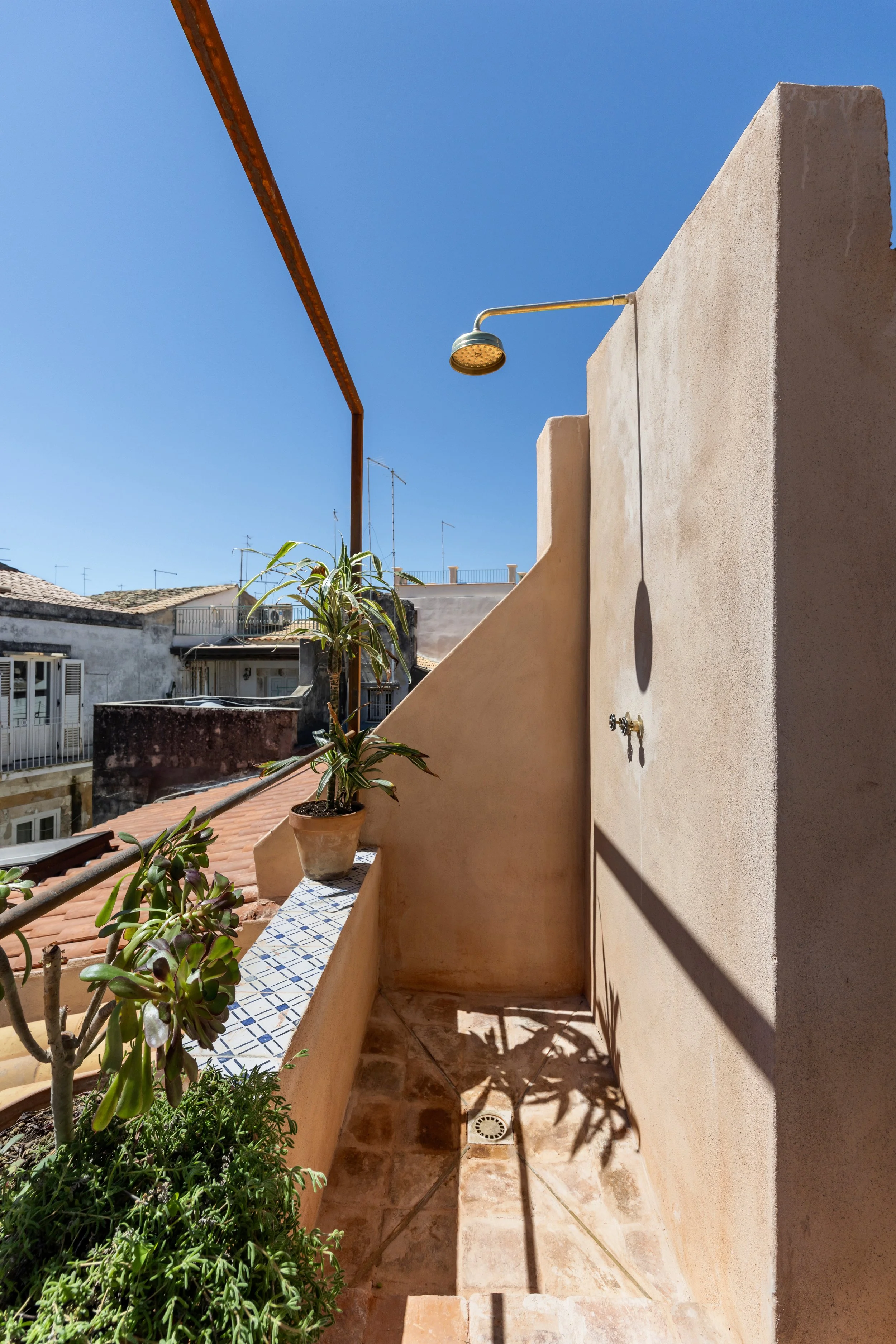 A small outdoor balcony with a faucet, potted plants, an outdoor shower head, and a railing. The sky is clear and sunny.