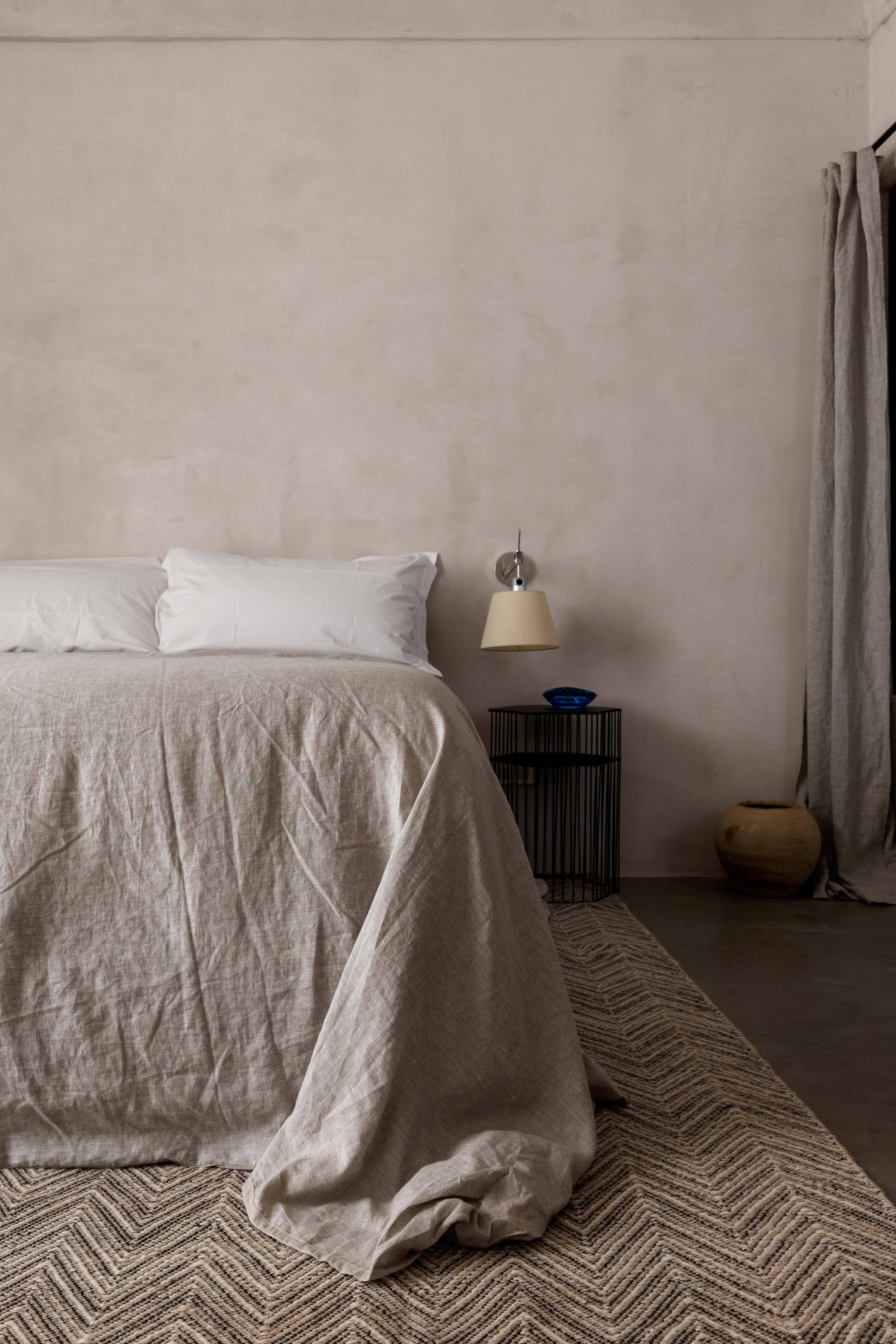 A bedroom with a bed covered in beige linen, two white pillows, a black side table with a blue bowl and a beige lamp, a textured beige and black rug, a ceramic pot on the floor, and beige curtains against a plain beige wall.
