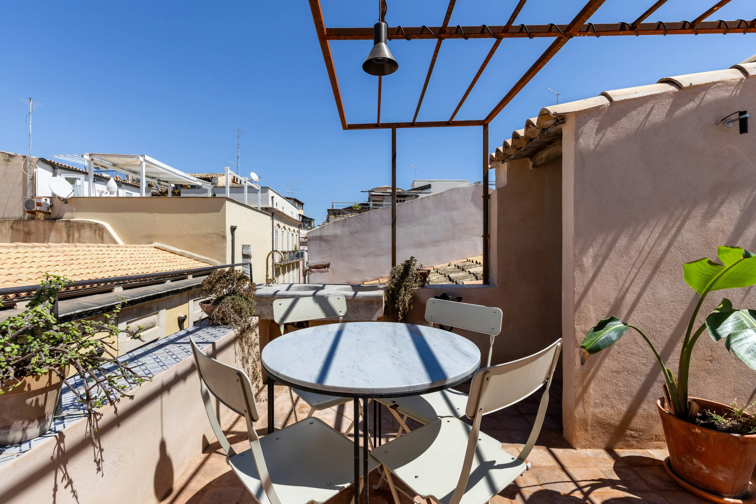 A rooftop balcony with a round white marble table and four white chairs, potted plants, and a black outdoor light fixture, with neighboring buildings and a clear blue sky in the background.