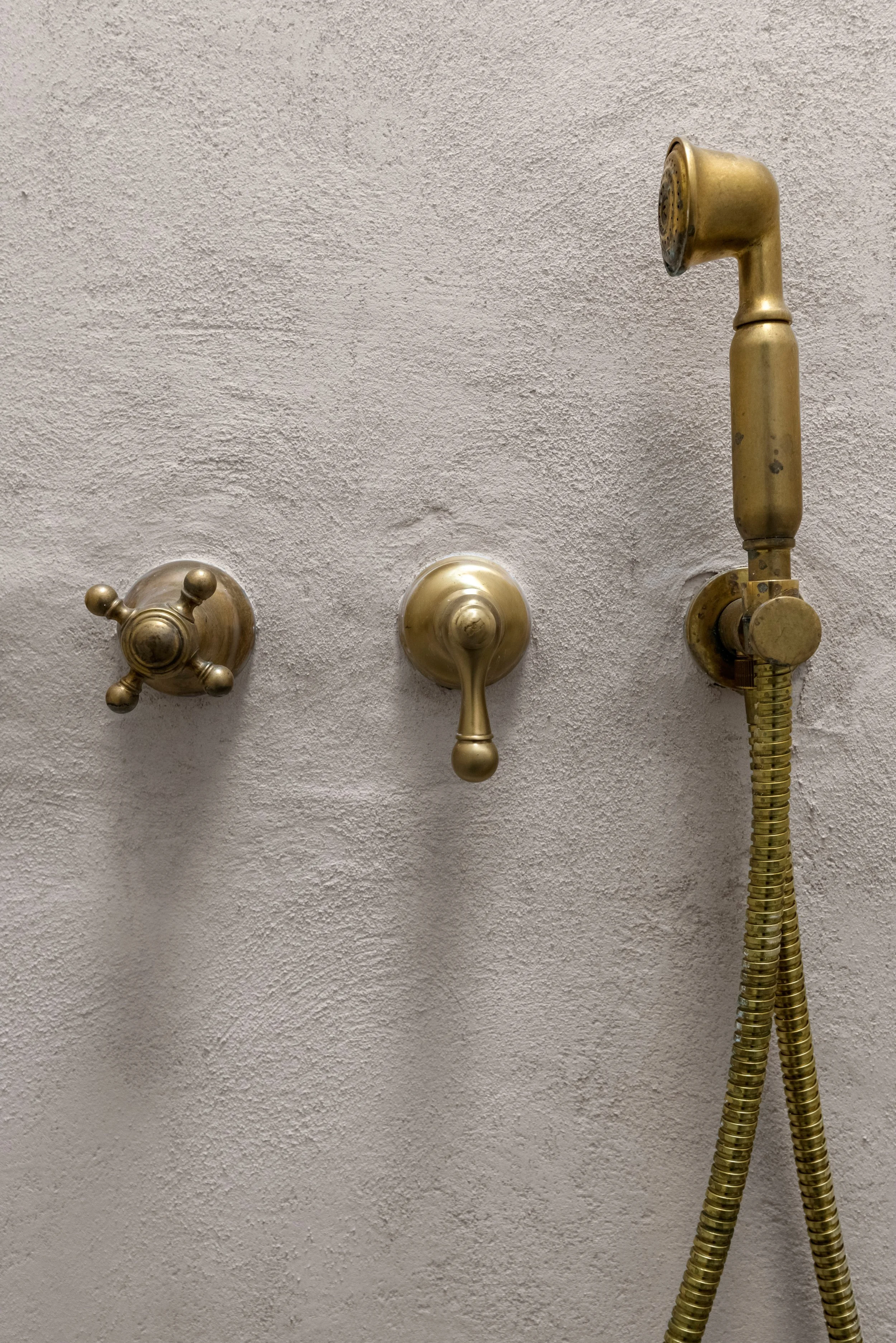 A wall with three vintage brass shower fixtures, including a handheld showerhead and two knobs for controlling water flow and temperature.