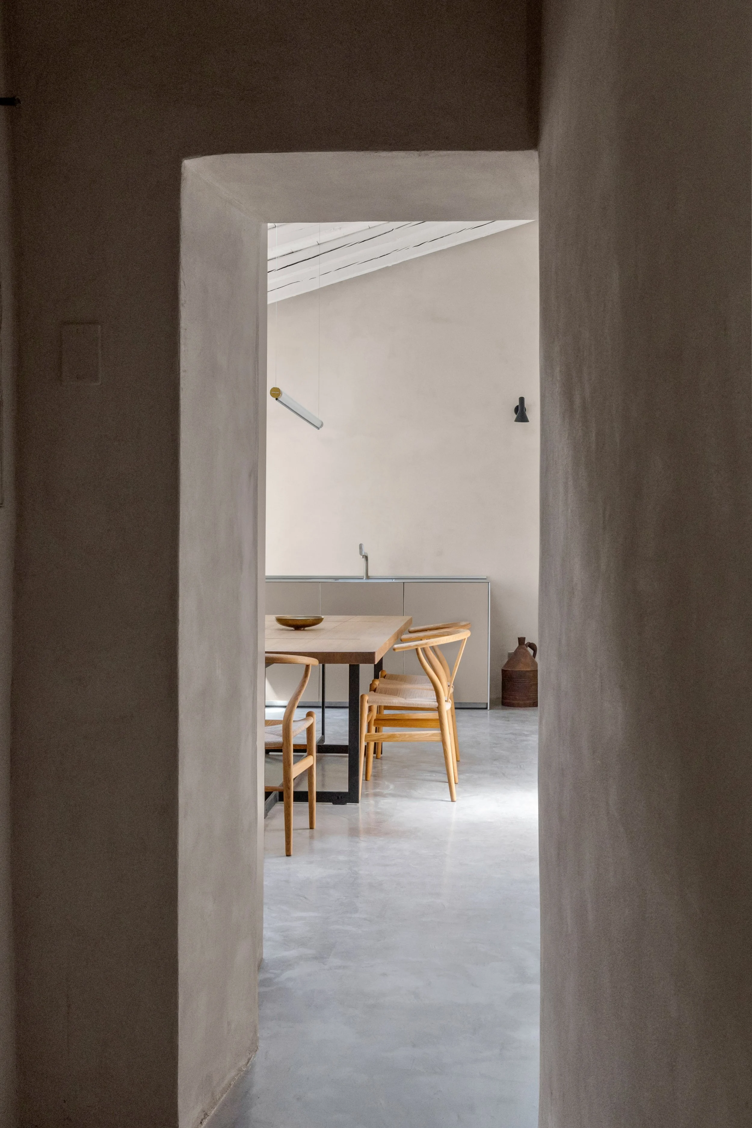 View through a doorway into a minimalist dining area with a wooden table, four wooden chairs, a sink, and a decorative vase against a beige wall.