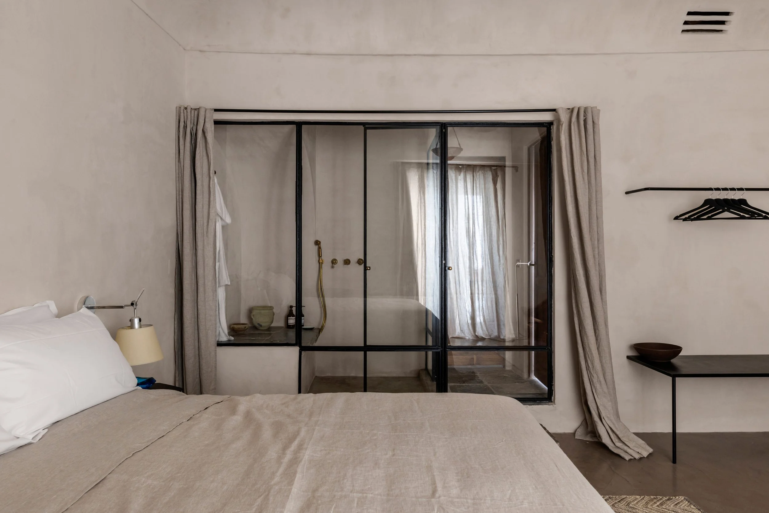 A bedroom with a beige bedspread, white pillows, a wall-mounted lamp, a small side table with a lamp, and a glass partition leading to a bathroom with a shower and curtain, and a curtain on the window in the background.