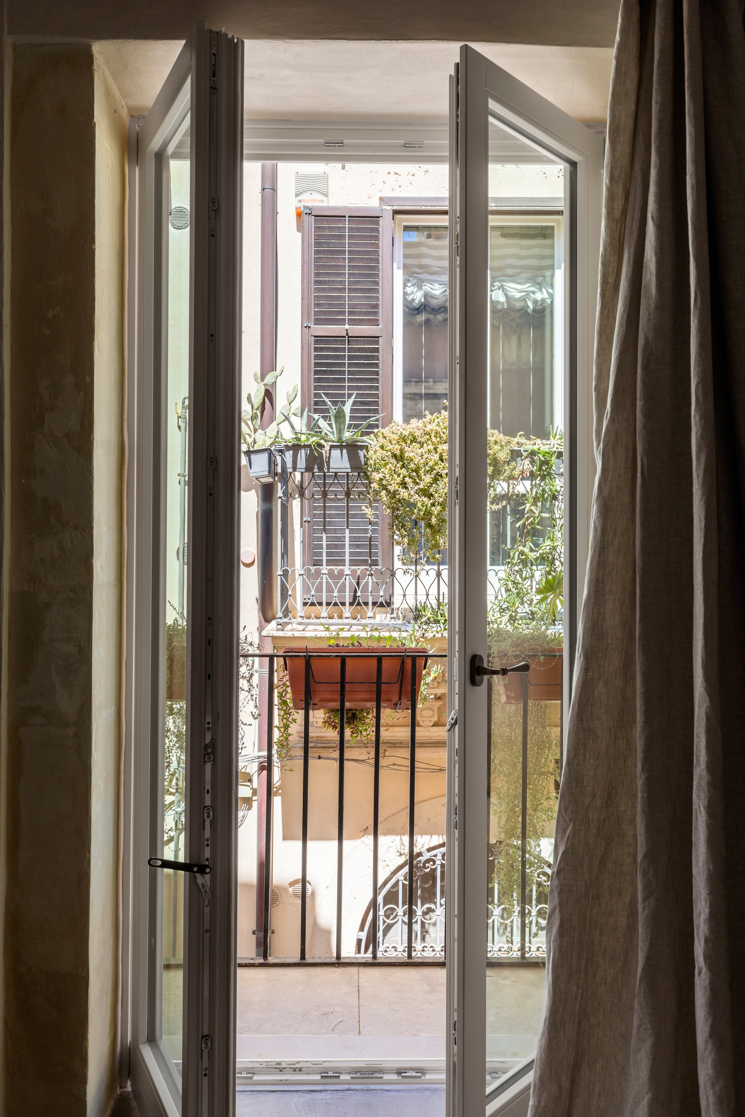 Open glass door leading to a small balcony with potted plants and a black metal railing, overlooking neighboring buildings with window shutters.