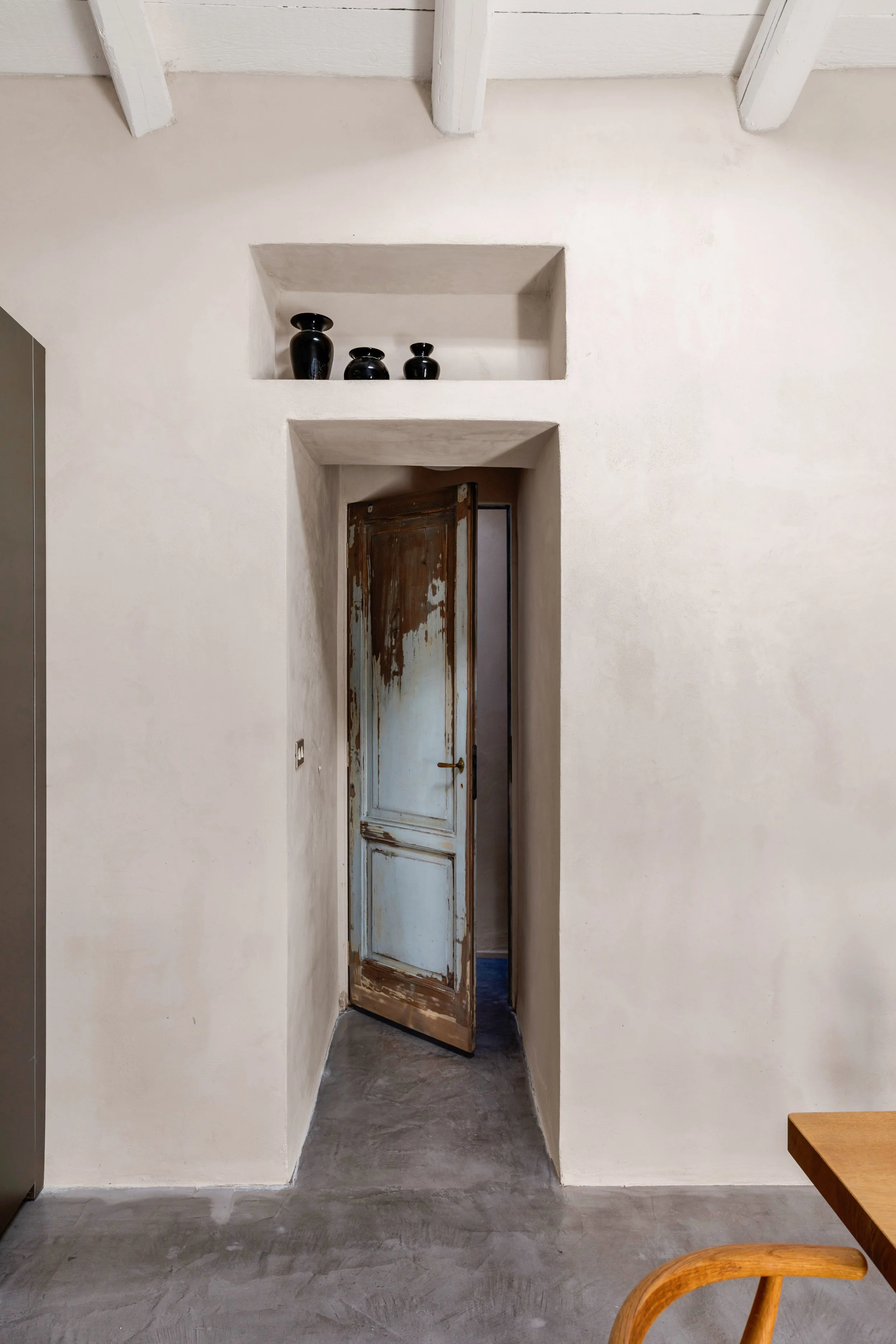 A worn wooden door slightly open in a white plaster wall, with decorative black vases on a built-in shelf above.