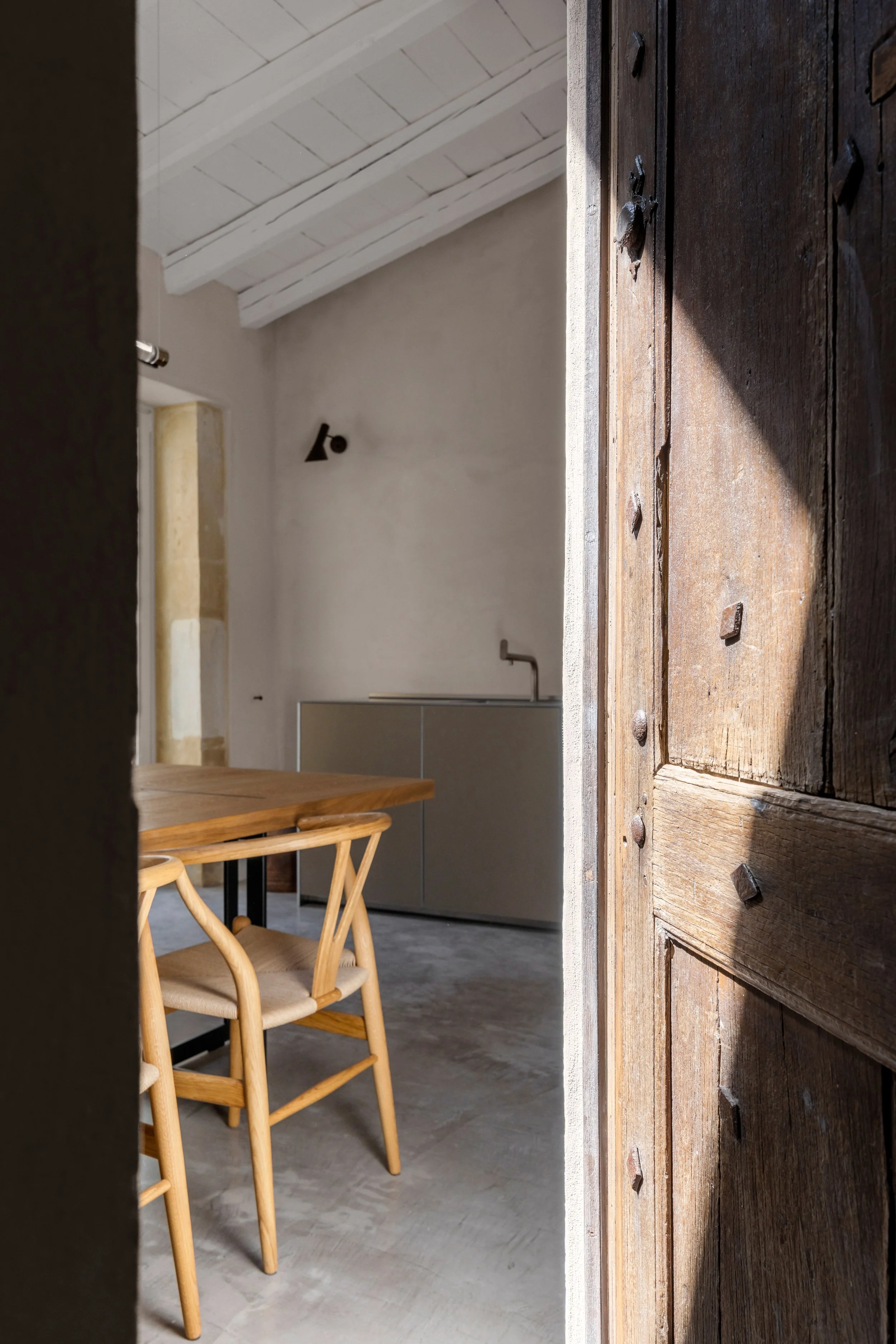 View through a slightly open rustic wooden door into a modern minimalist room with a wooden dining table, light wooden chairs, a gray cabinet, and a white ceiling with exposed beams.