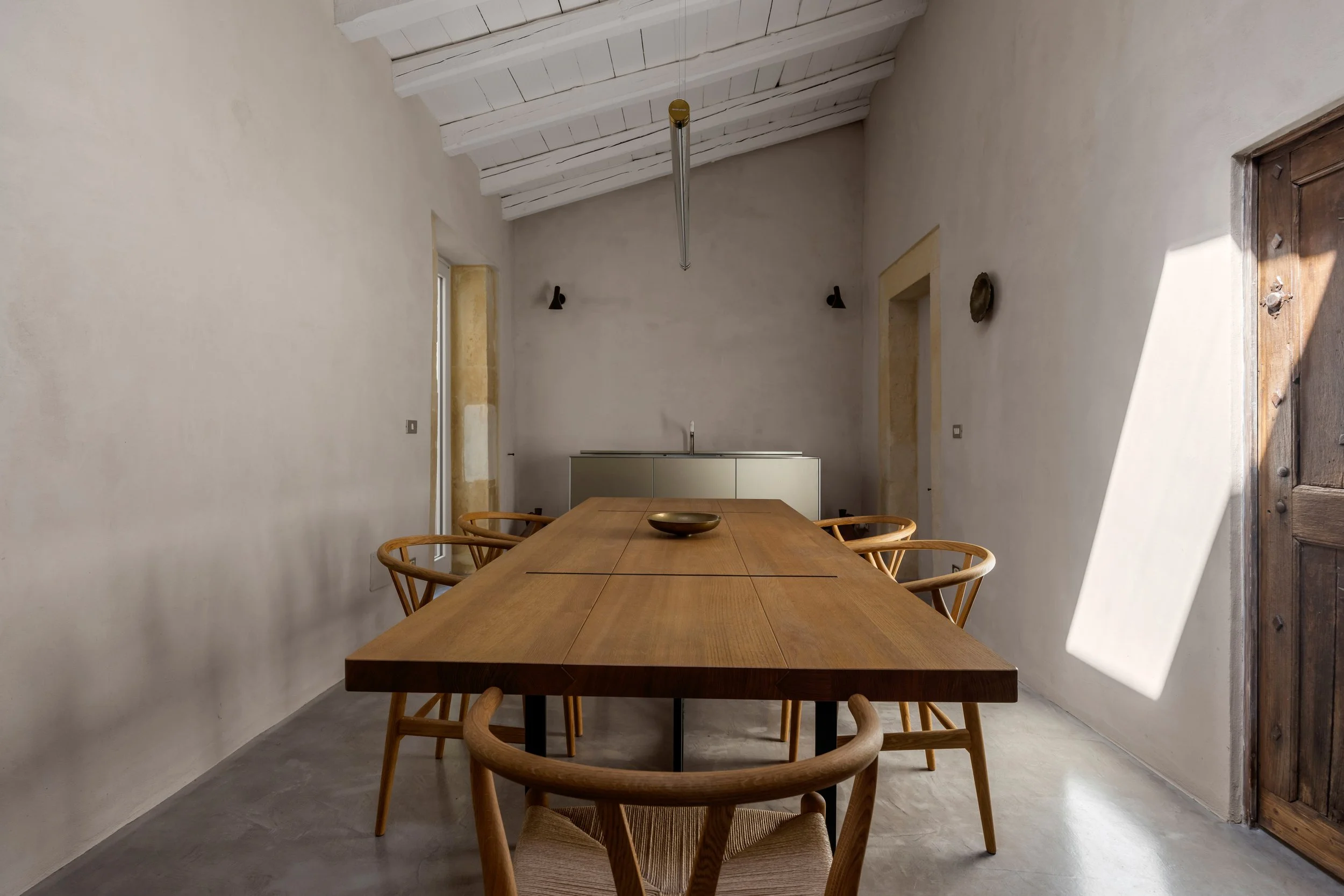 Minimalist dining room with a large wooden table, eight wooden chairs, a small bowl on the table, a ceiling with white exposed beams, and sunlight streaming through a window casting shadow on the wall.
