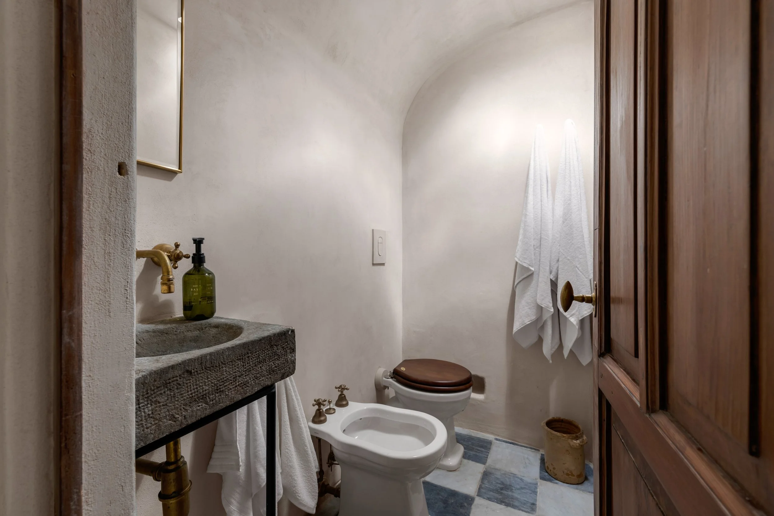 Small bathroom with a rustic stone sink, a toilet with a wood seat, a wooden cabinet, and white towels hanging on the wall.