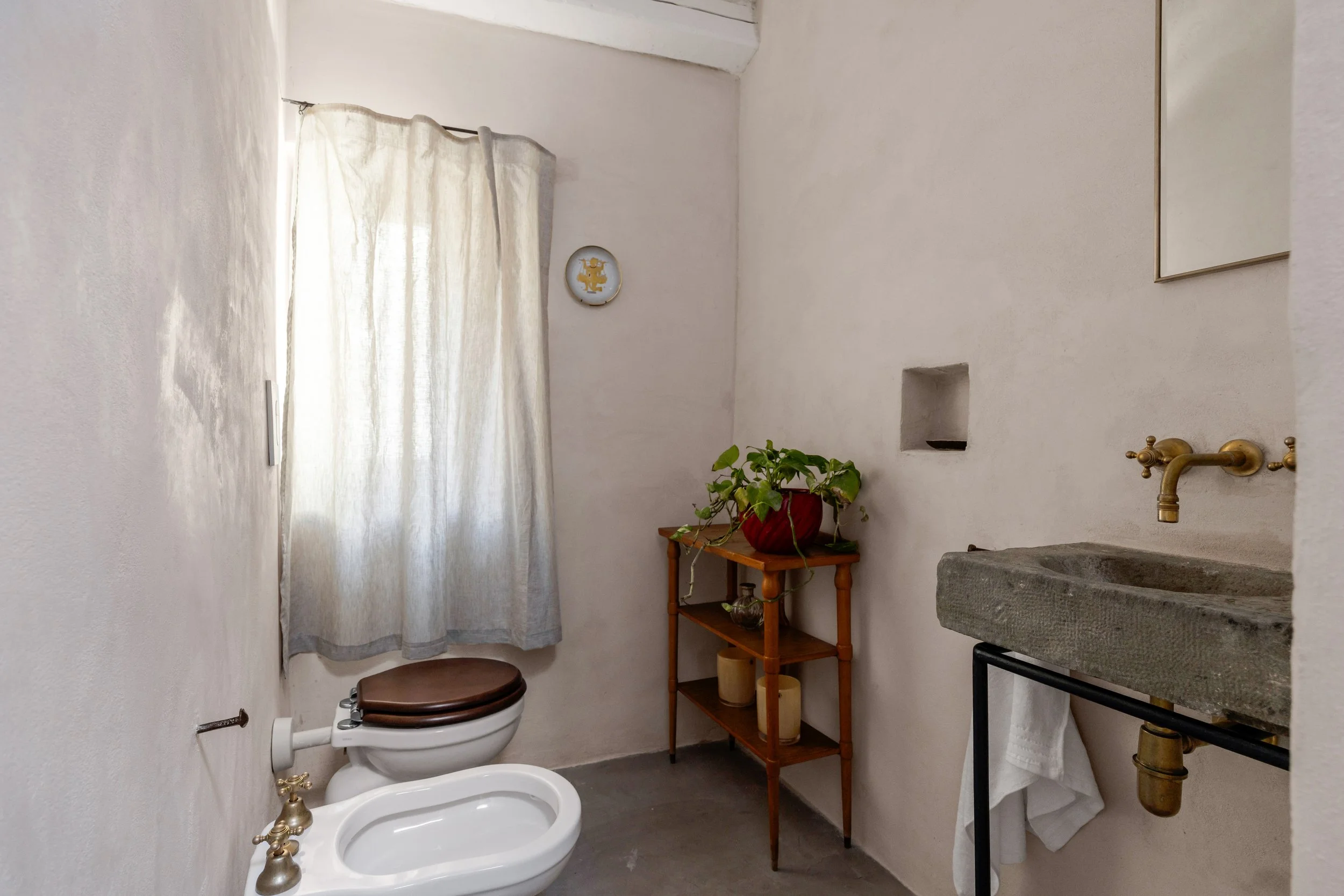 A minimalist bathroom with a toilet, potted plant on a wooden stand, a stone sink with brass fittings, and a small towel hanging beneath the sink. There is a window with sheer curtains and a decorative plate on the wall.