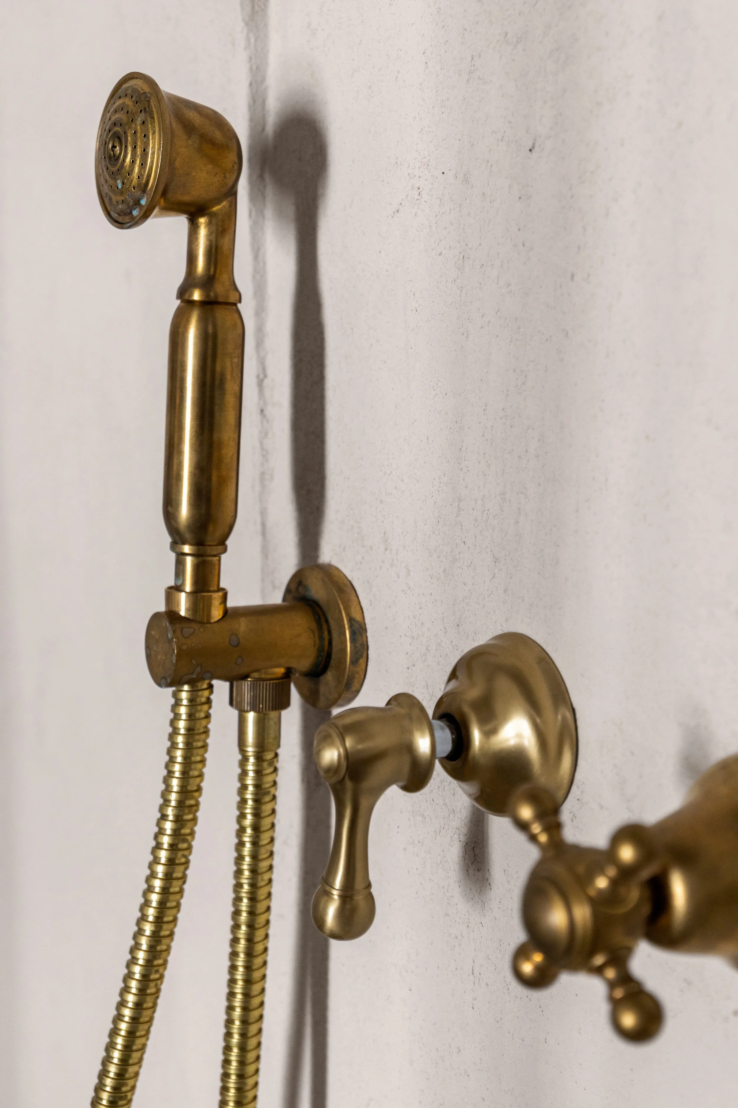 Close-up of vintage brass bathroom fixtures on a gray wall, including a handheld showerhead with a hose and separate faucet handles.