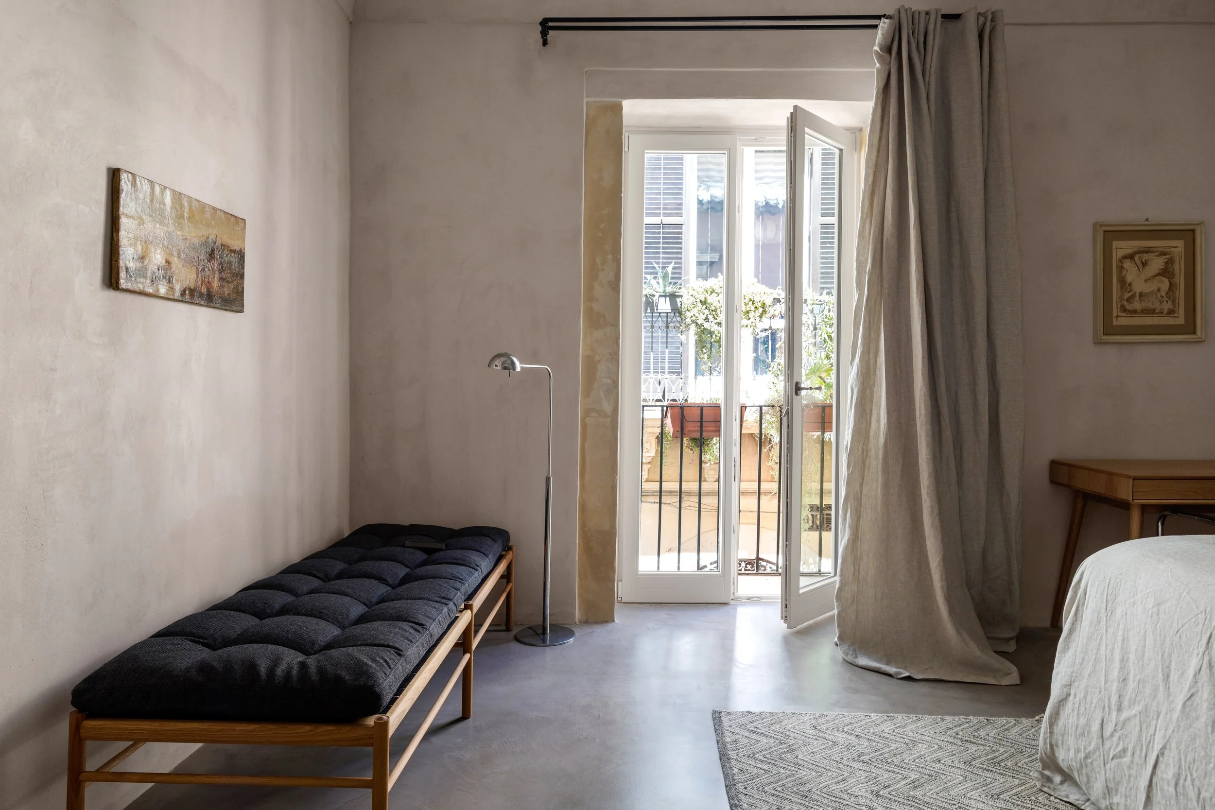 A minimalist bedroom with an open balcony door. It features a black cushioned bench, a metallic floor lamp, a wooden nightstand, and a bed with a white bedspread. Light beige curtains are partially drawn to the side of the door, allowing sunlight to illuminate the room.