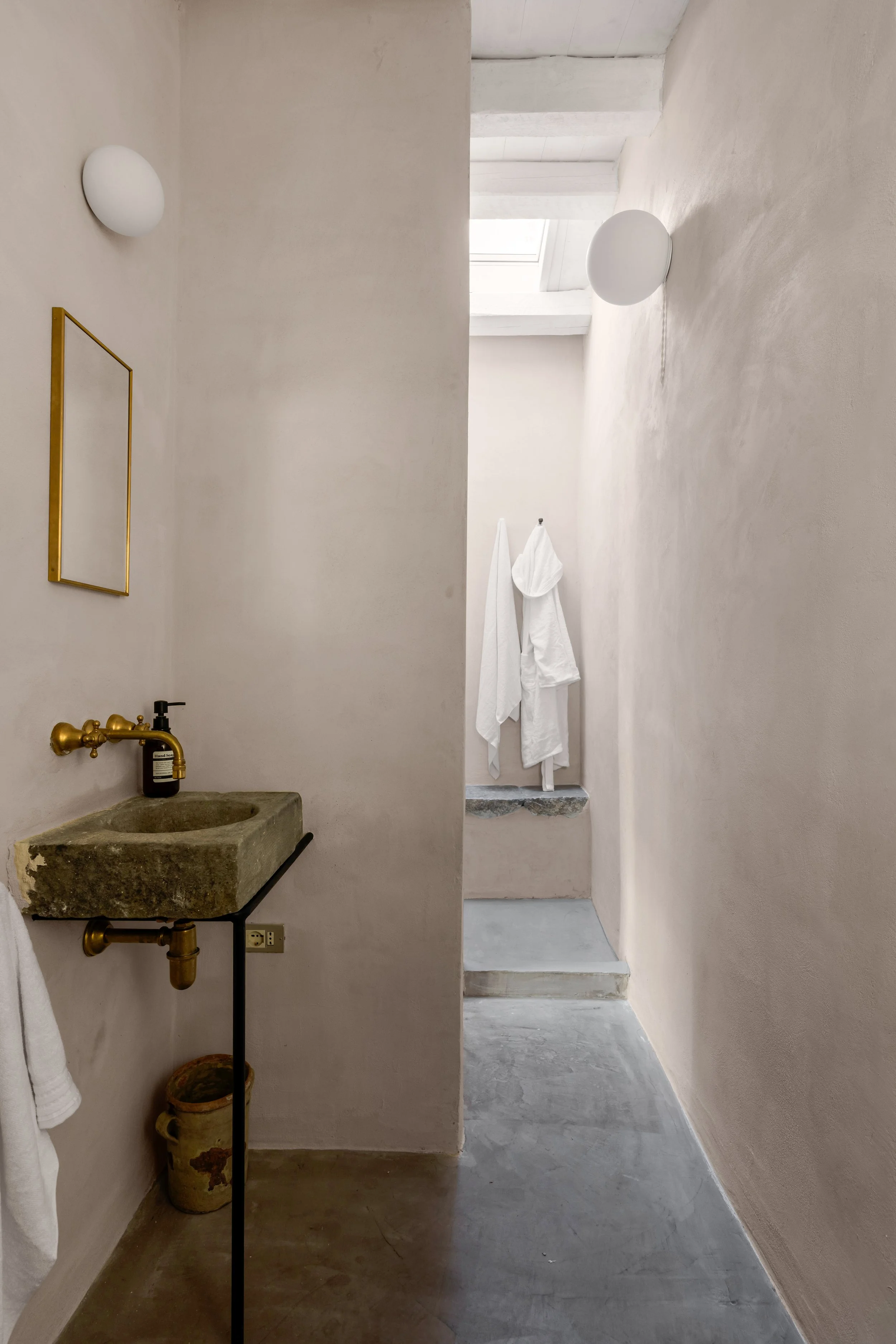 Minimalist bathroom with concrete sink, gold fixtures, and white towels hanging in a bright, airy space with a high ceiling and skylight.