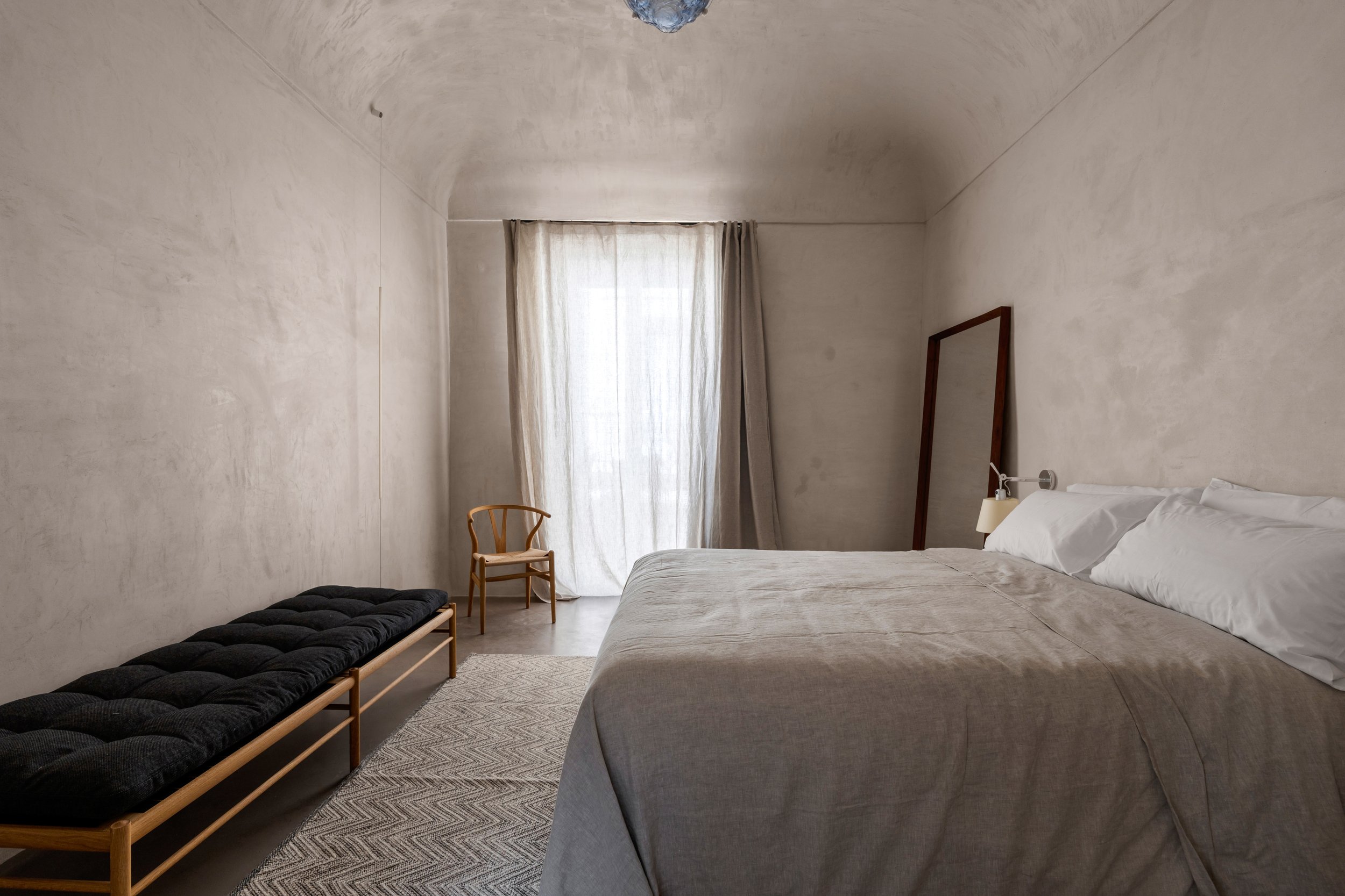 Minimalist bedroom with a large bed, a bench, a chair, a mirror leaning against the wall, and light-colored curtains on a window.